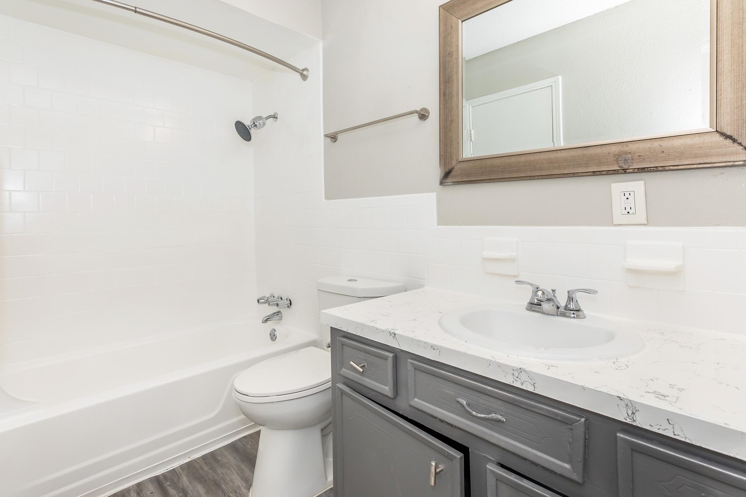 A modern bathroom featuring a white bathtub with a showerhead, a toilet, and a gray vanity with a marble countertop. A large mirror is above the sink, and the walls are painted light gray. The flooring is a wood-style laminate.