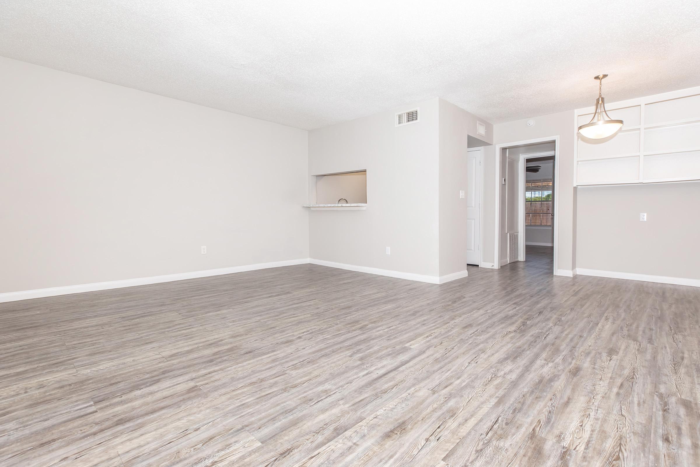 A spacious, empty living room featuring light-colored walls, a smooth wood-like floor, and a ceiling light fixture. There is an open layout with a doorway leading to another room and a small bar area open to the kitchen. The ambiance is bright and inviting, perfect for personal customization.