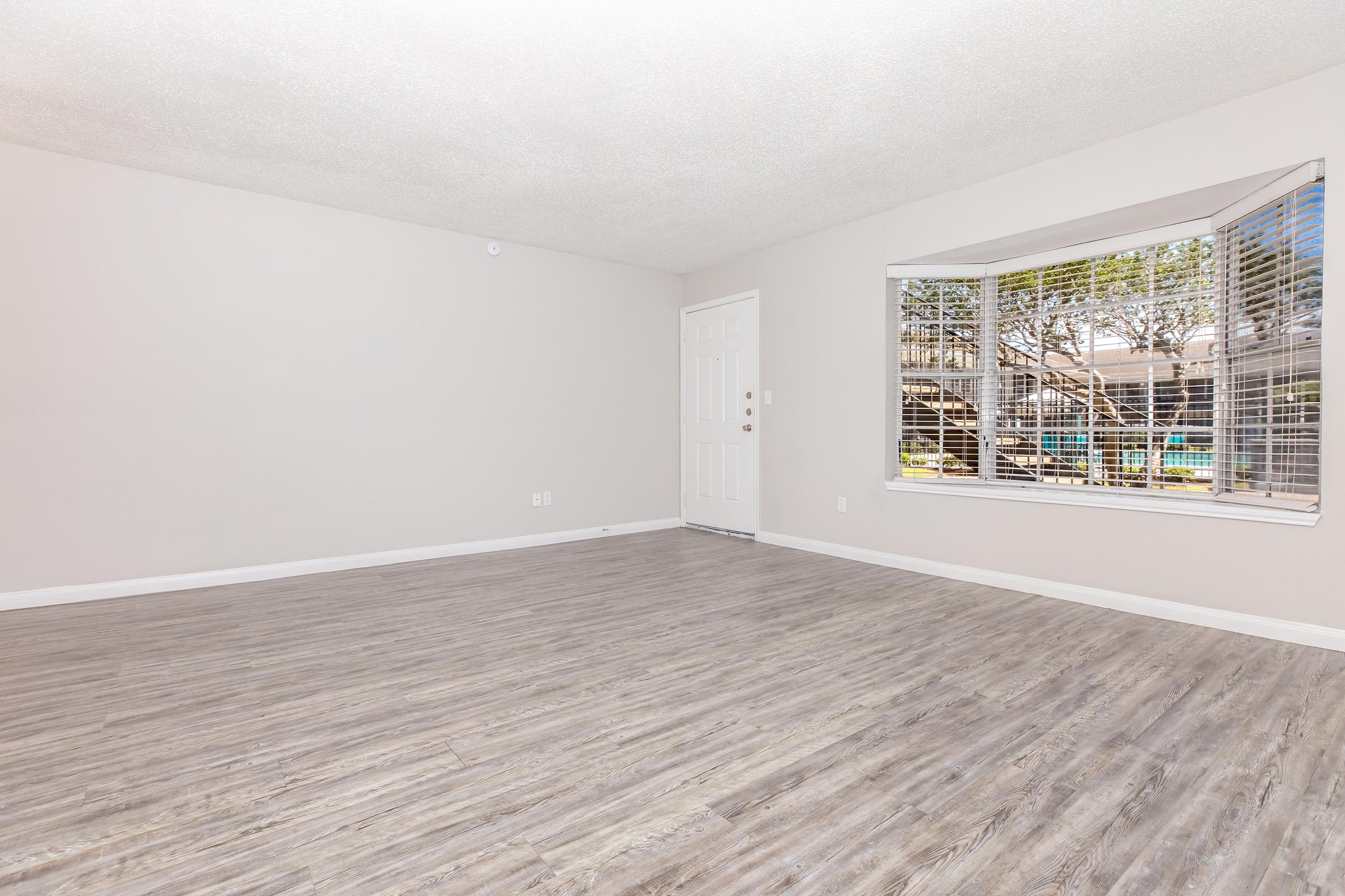 Empty room featuring light-colored walls and a textured ceiling. The floor has a modern wood-like finish. A large window with multiple panes allows natural light to enter, and there's a door leading outside. The room is spacious and uncluttered, providing a blank canvas for decoration.