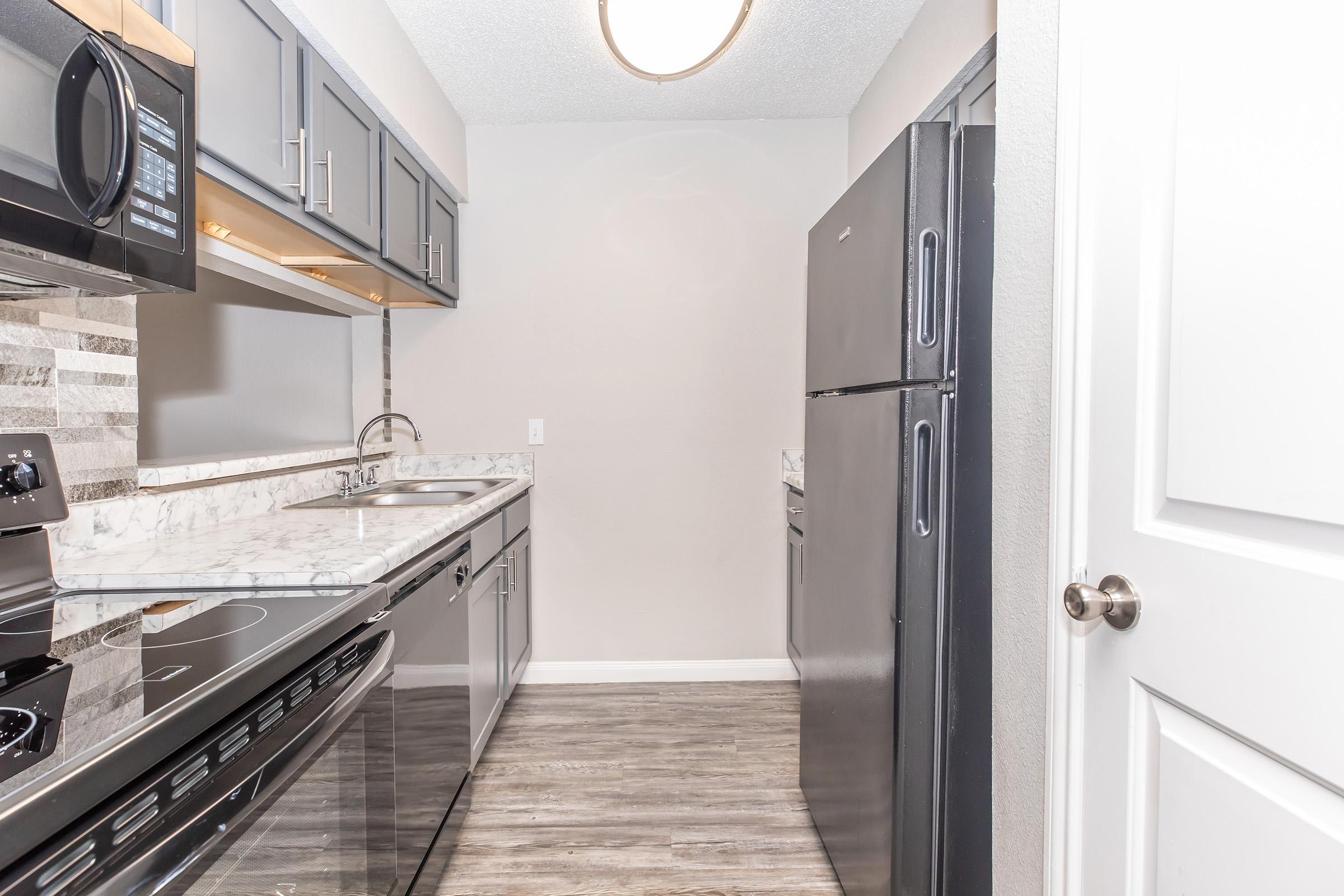 Modern kitchen with gray cabinets, stainless steel appliances, and a marble countertop. Features an electric stove, microwave, sink, and a black refrigerator. The flooring is wood-like laminate, and the space is well-lit with a ceiling light and neutral-colored walls.
