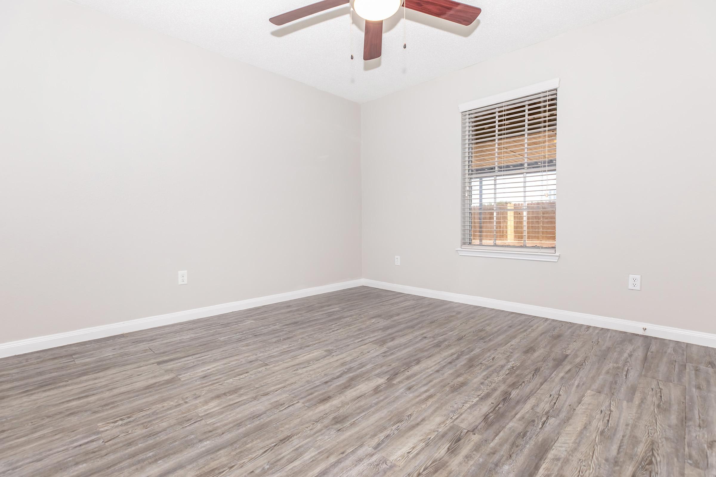 Empty room with light gray walls and wood-like flooring. A ceiling fan with wooden blades is installed. There is a window with blinds allowing natural light to enter, and the space appears clean and minimalistic, ready for furnishing.
