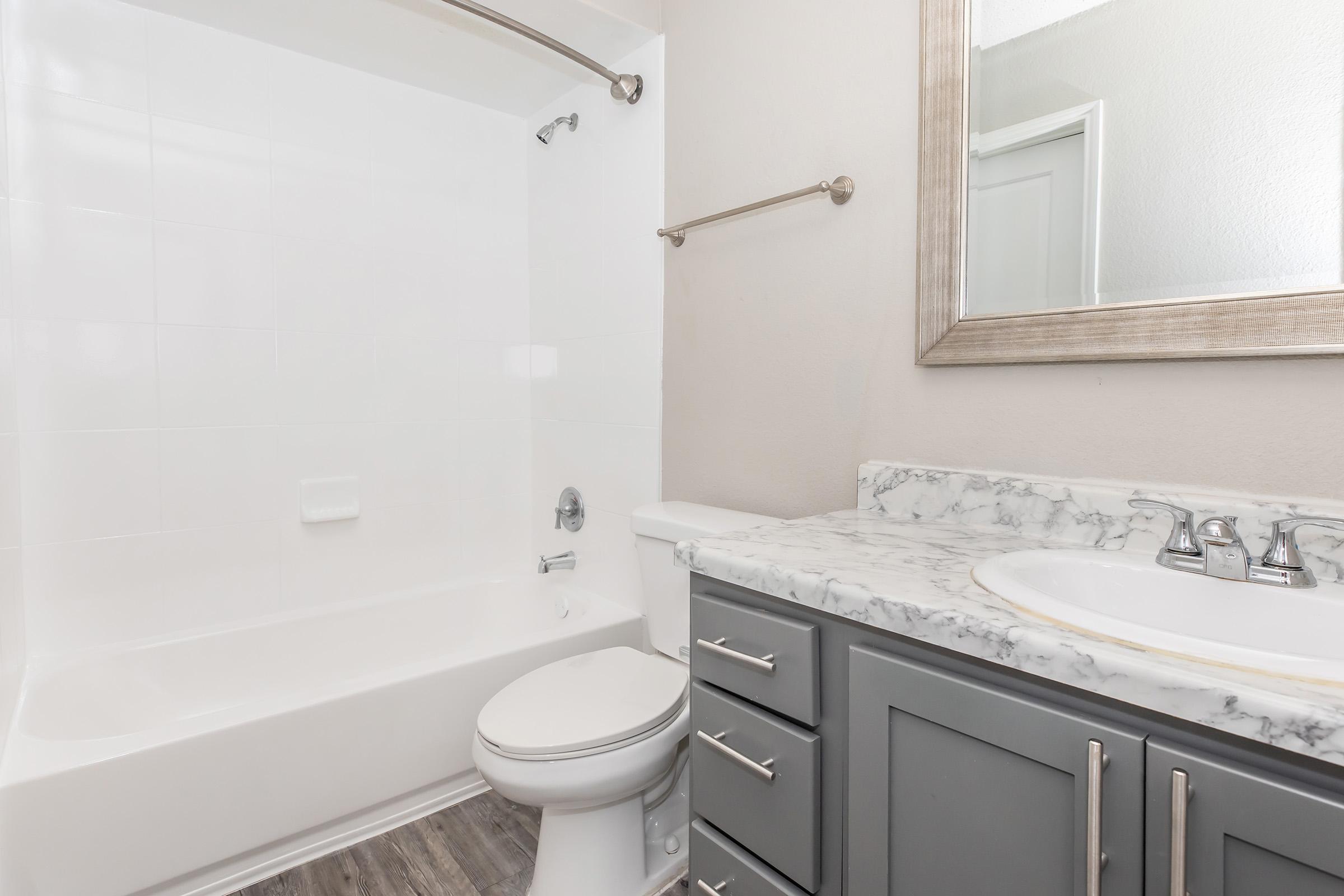 A clean, modern bathroom featuring a white bathtub and shower combination, a marble countertop with a sink, a gray cabinet with drawers, and a large mirror above the sink. The walls are a neutral color, and the flooring is a light wood look. Natural light illuminates the space.