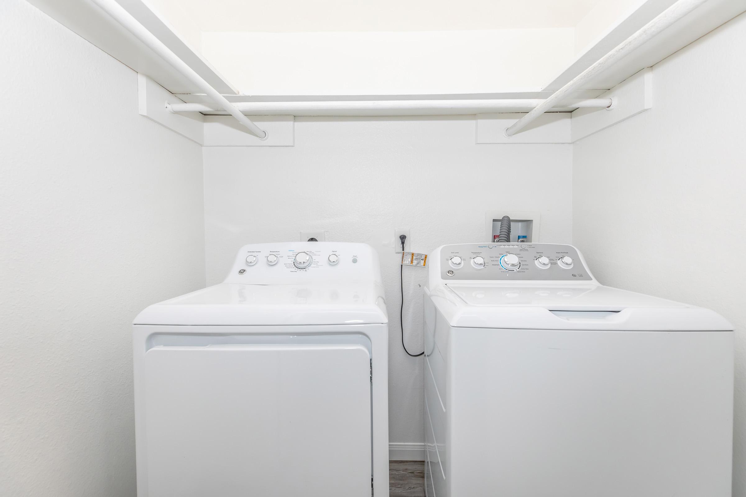 A clean laundry room featuring a stacked pair of white washing machines and dryers, positioned side by side against a light-colored wall. The space is well-organized with minimal decorations, showcasing a functional area for laundry tasks.