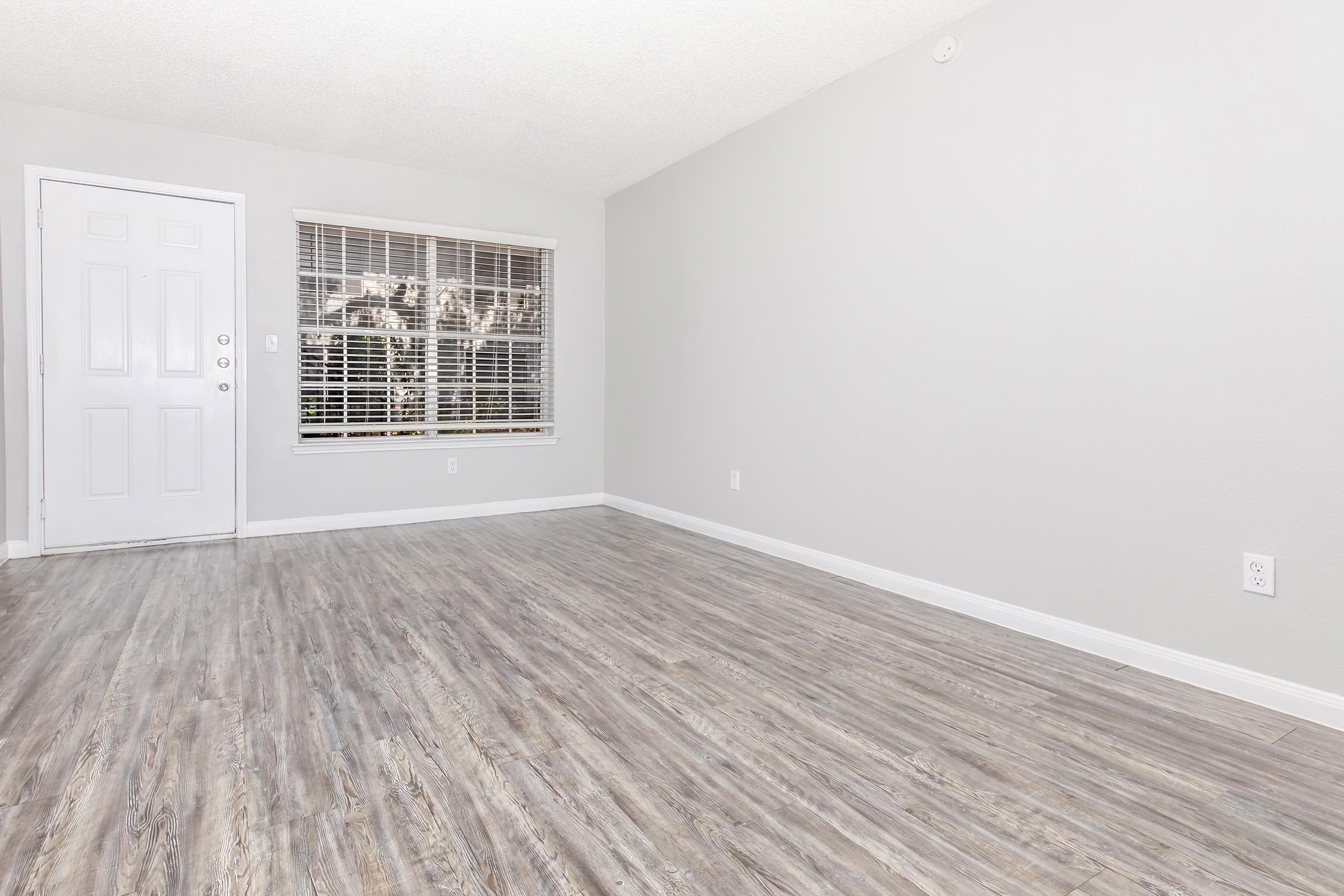 A spacious, empty living room with light gray walls and wooden-style flooring. There's a front door on the left and a large window with white blinds allowing natural light, creating a bright and airy atmosphere. The room is ready for furniture and personalization.