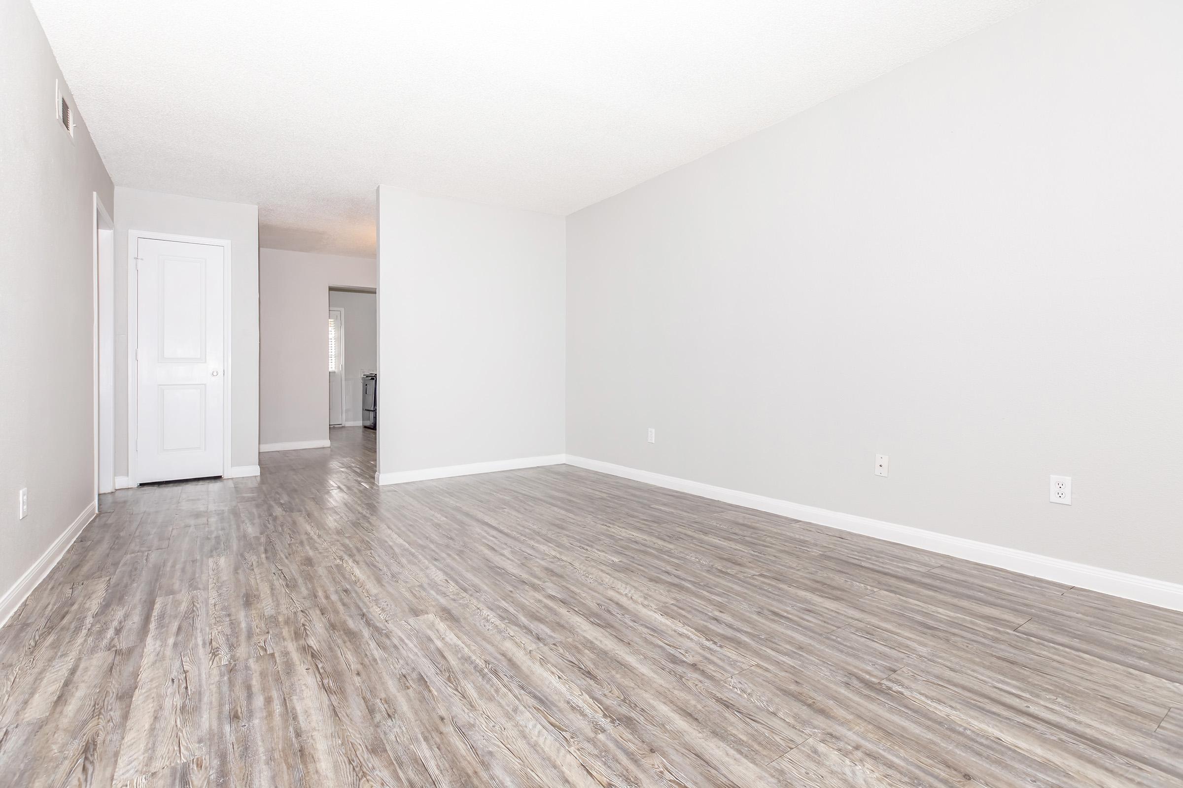 A spacious, empty room with light gray walls and a modern light-colored laminate floor. There's a white door on the left and an open space leading to another room in the background. Natural light fills the space, creating a bright and inviting atmosphere.