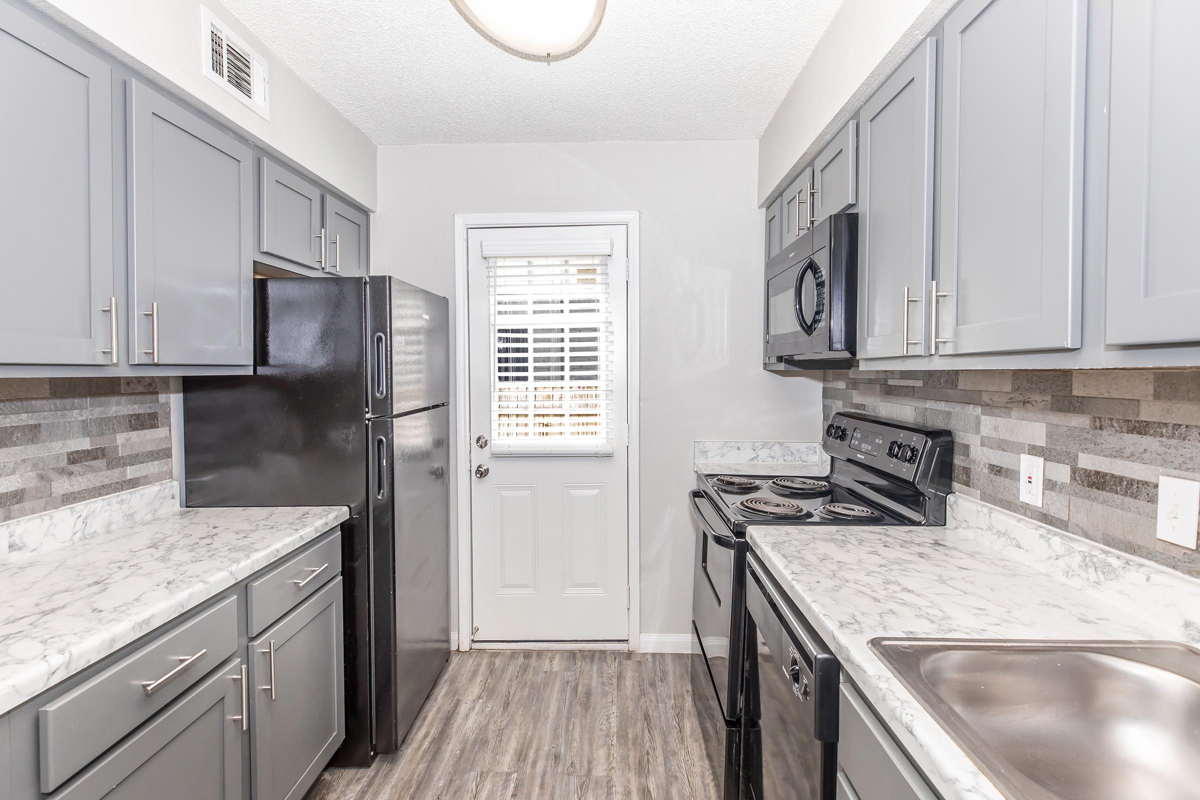 A modern kitchen featuring gray cabinetry, marble countertops, and stainless steel appliances. The layout includes a refrigerator, stove, and sink, with gray and white stone backsplash. A door with a window leads outside, and the flooring is made of wood-like laminate.