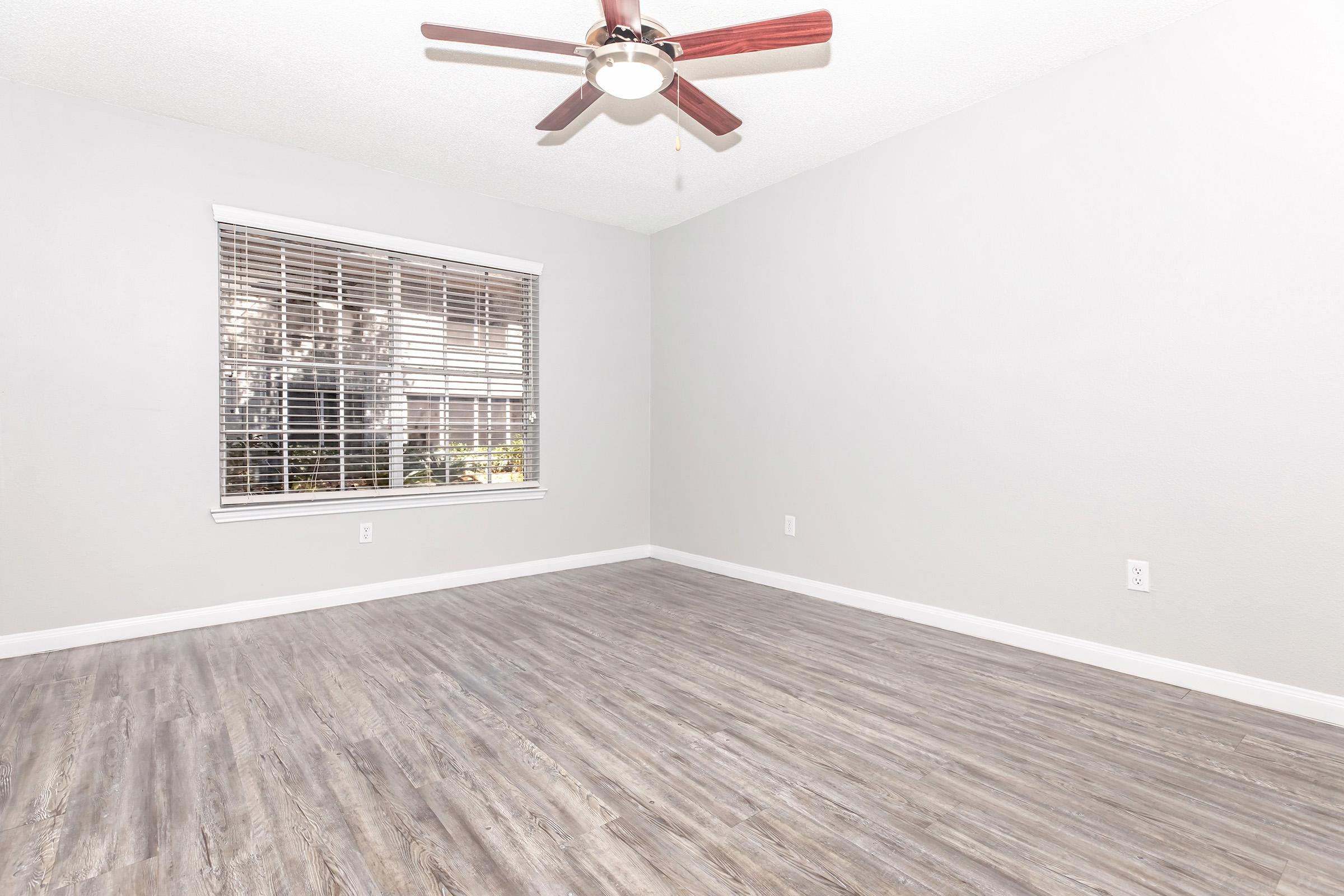 Empty room with light gray walls and a ceiling fan. The floor is made of wood-like laminate, and there is a window with closed blinds allowing natural light. The space is clean and spacious, suitable for various uses like a living area or office.