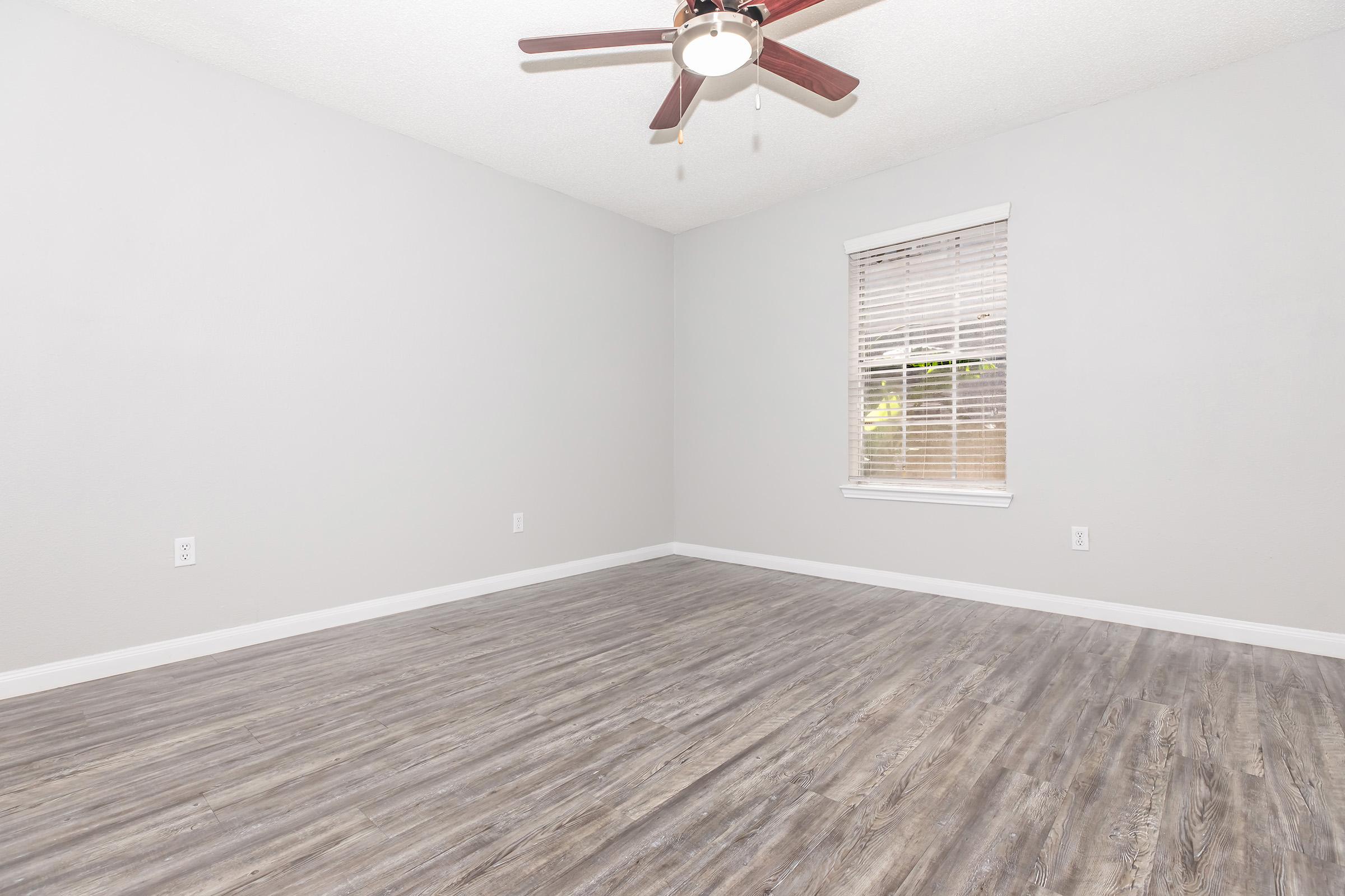 A spacious, empty room with light gray walls and a ceiling fan. The flooring is a wood-look laminate in a medium tone. A window with blinds allows natural light to enter, highlighting the clean and minimalistic atmosphere of the space.