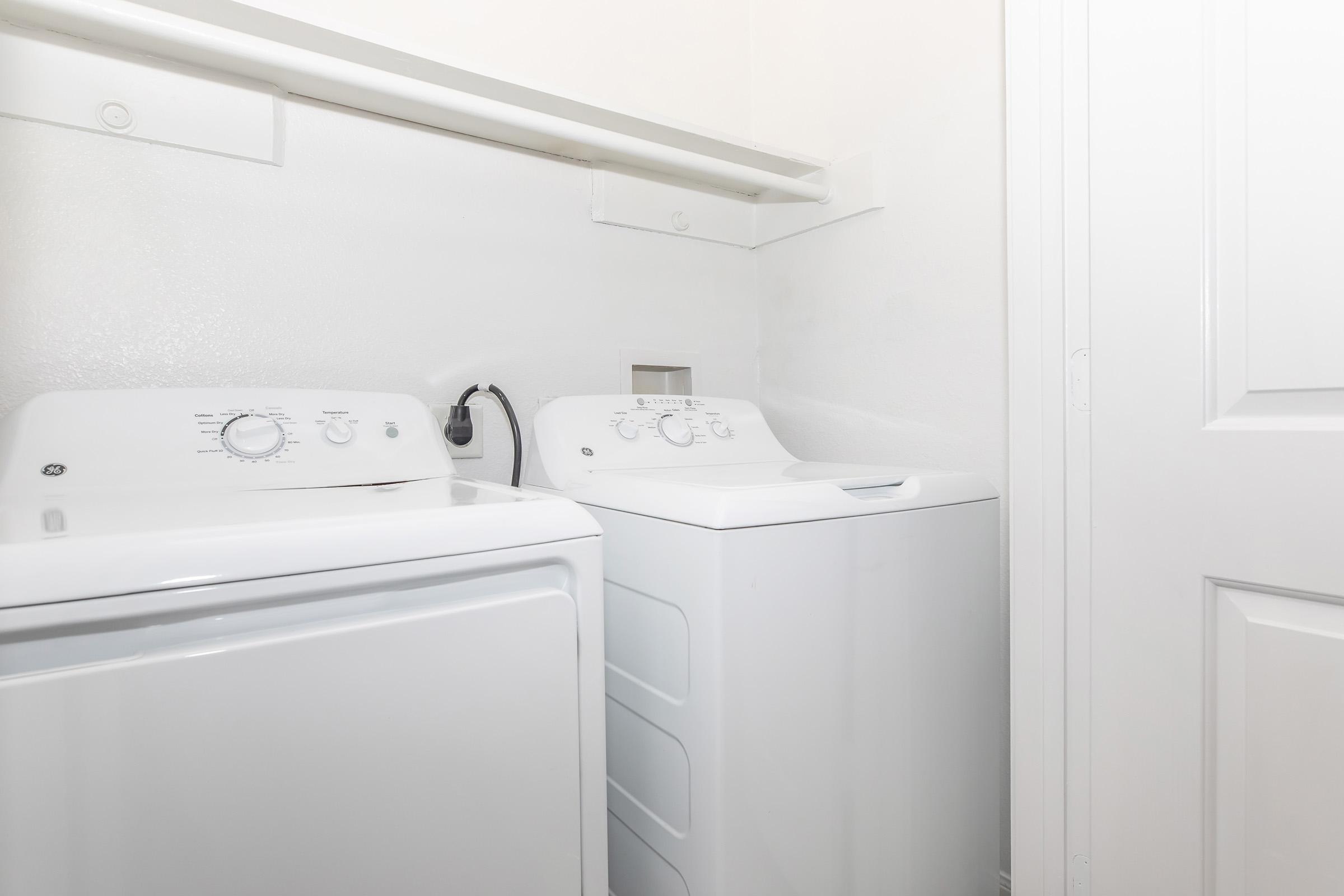 A neatly organized laundry area featuring a white front-loading washing machine on the left and a white top-loading washing machine on the right, both against a plain wall. A shelf is mounted above the machines for storing laundry supplies. The setting is bright and clean.