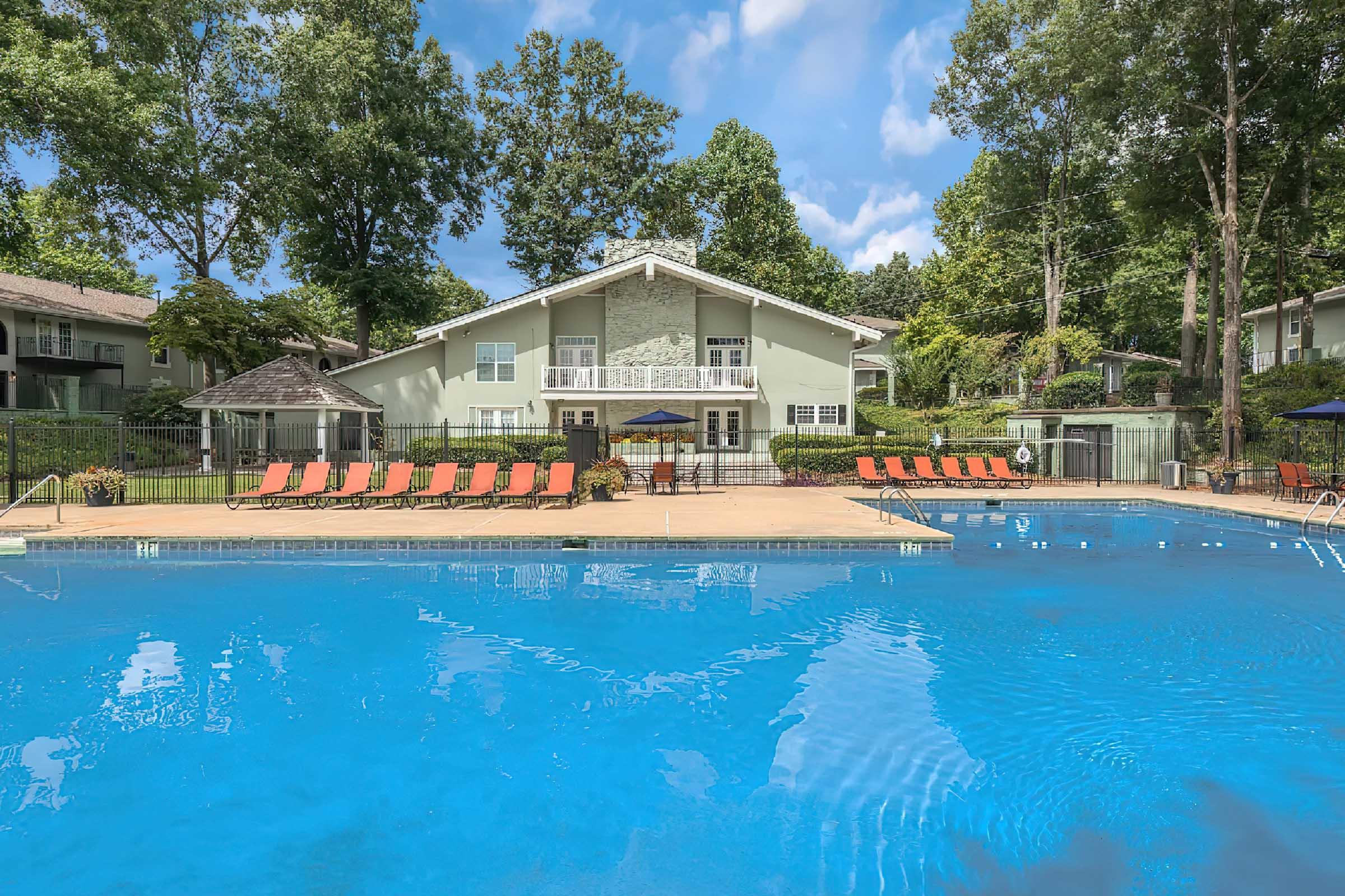 A bright outdoor swimming pool surrounded by lounge chairs and greenery, with a multi-story building in the background. The scene is set on a sunny day, highlighting a relaxing atmosphere ideal for leisure and enjoyment.