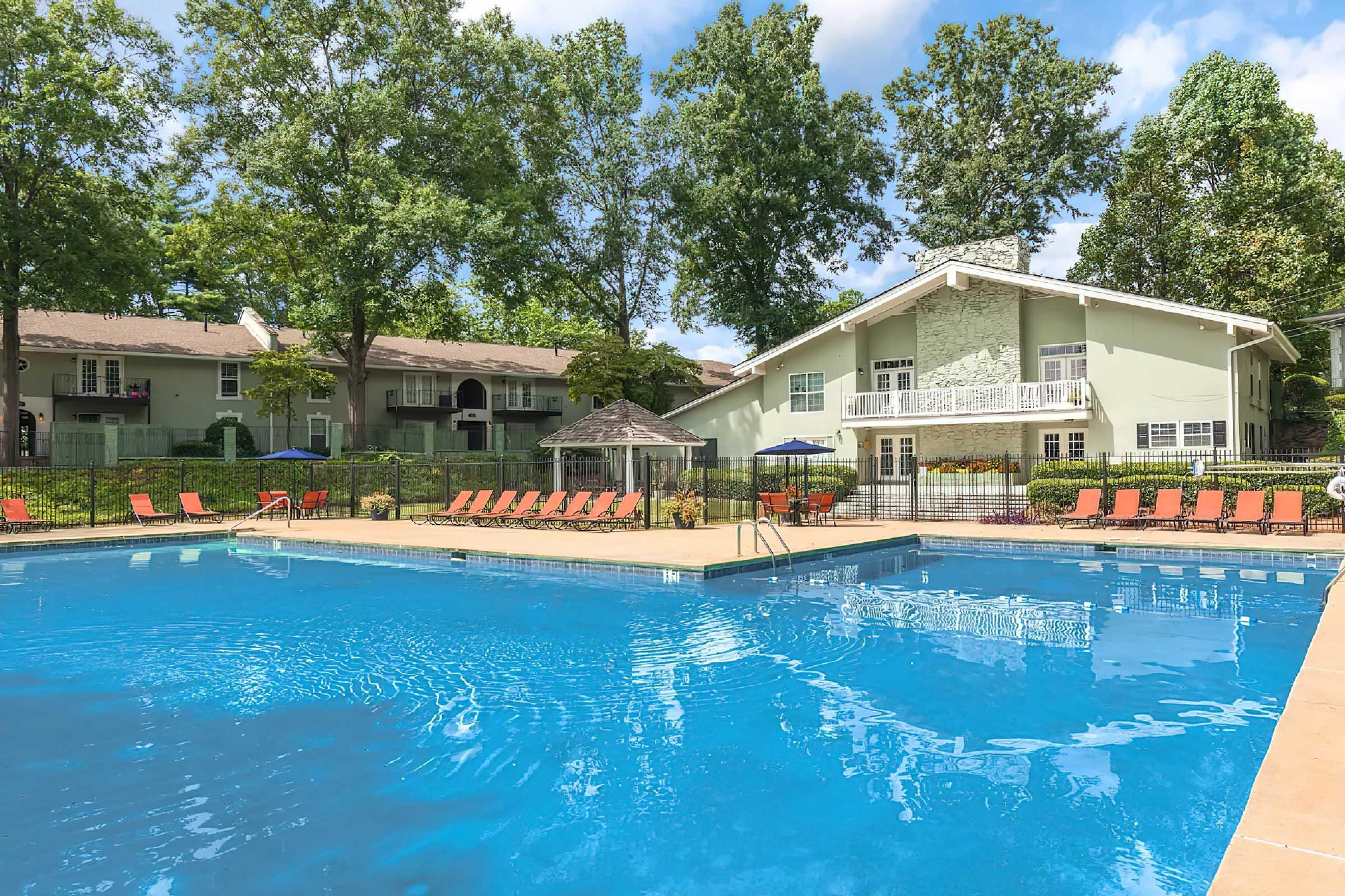 A sparkling blue swimming pool surrounded by lounge chairs and shaded by trees. In the background, a light green building with balconies overlooks the pool area, along with a gazebo. Bright blue skies with fluffy white clouds create a sunny atmosphere in the setting.
