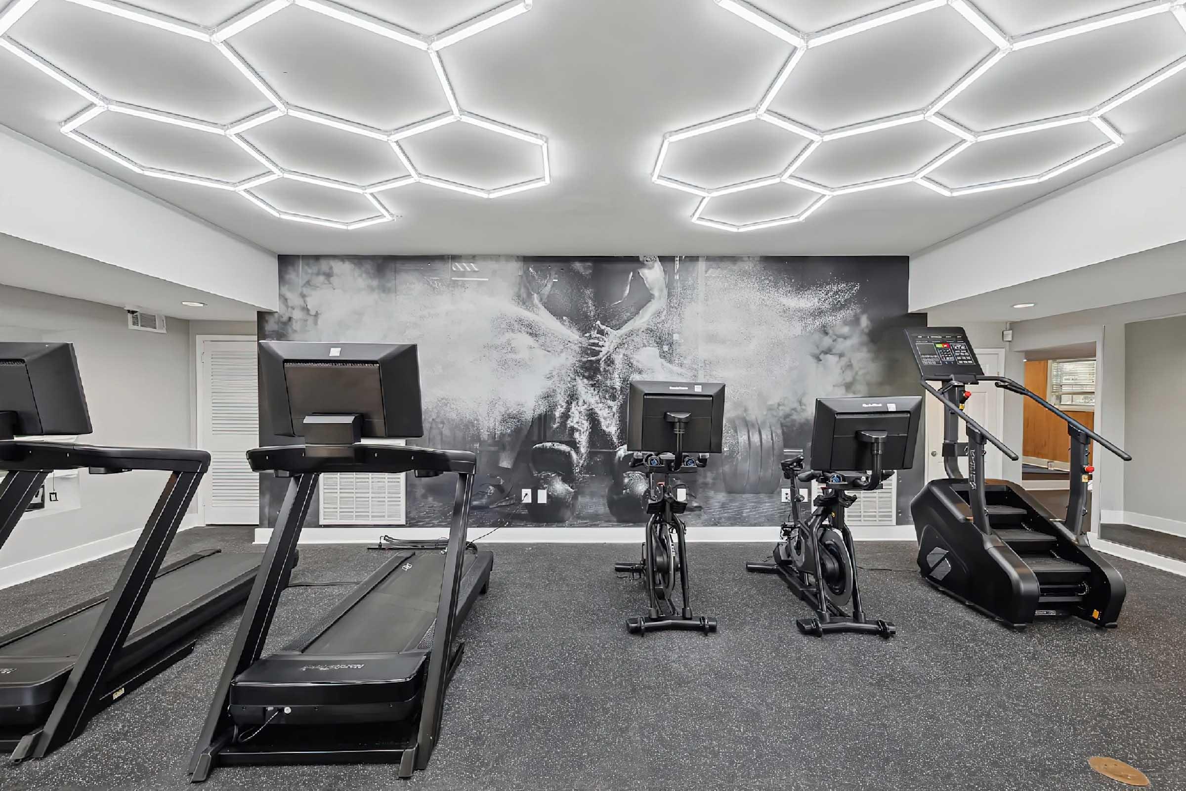 A modern fitness room featuring several treadmills, elliptical machines, and stationary bikes. The space has a sleek design with hexagonal lighting fixtures and a large mural on the wall. The flooring is black rubber, creating a professional workout environment.