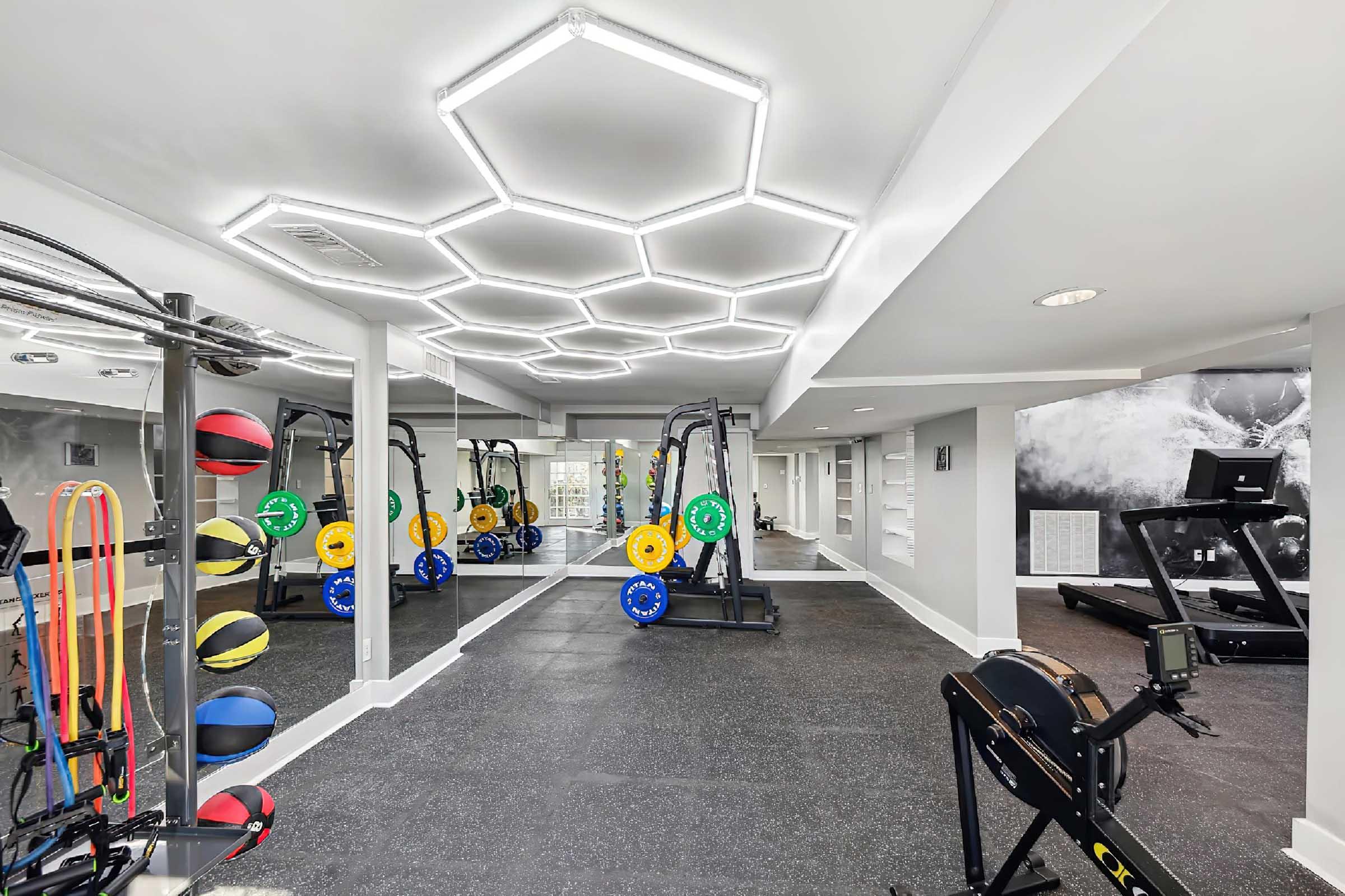 A modern gym interior featuring exercise equipment, including a rowing machine and free weights. Bright hexagonal ceiling lights illuminate the area, which also has colorful resistance bands and a treadmill. The flooring is dark, enhancing the spacious and contemporary feel of the workout space.