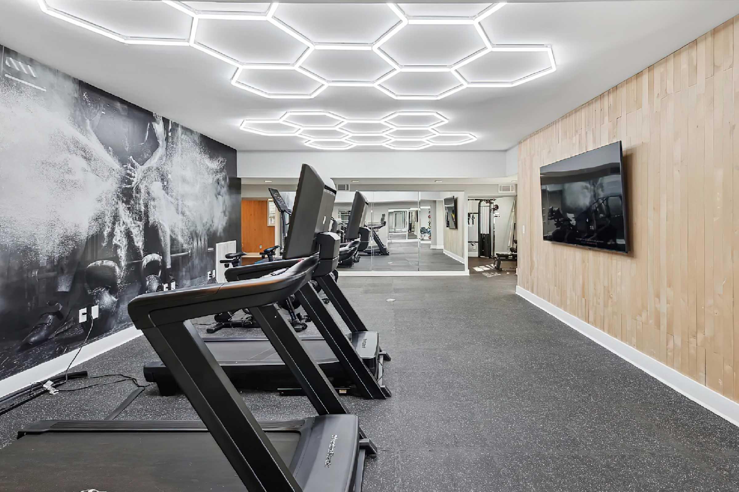 A modern gym interior featuring several treadmills aligned against a black and white wall mural. Bright hexagonal ceiling lights illuminate the space, which also includes a wall-mounted TV. The flooring is dark, and large mirrors reflect the equipment, creating an open and inviting workout environment.