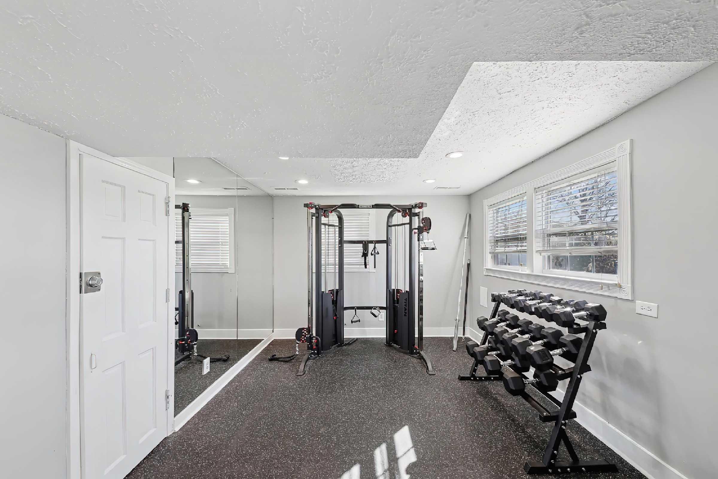 Interior view of a home gym featuring a cable machine, a rack of dumbbells, and mirrors. The room has gray walls, a textured ceiling, and natural light coming from windows. The flooring is dark rubber, suitable for exercise activities.