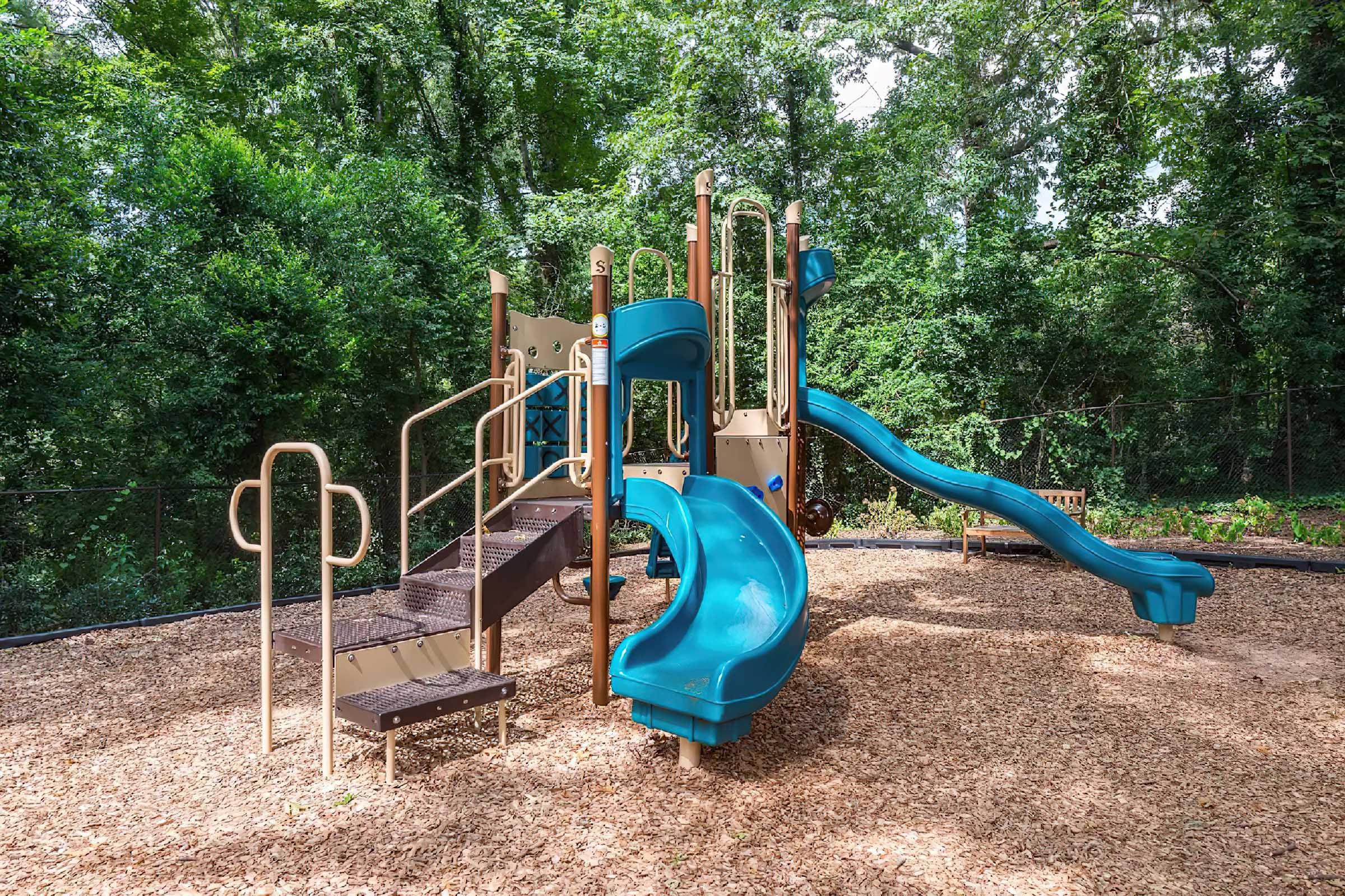 Playground with two blue slides, a climbing structure, and stairs, surrounded by trees and wood chips on the ground. The area appears to be well-maintained and inviting for children to play.