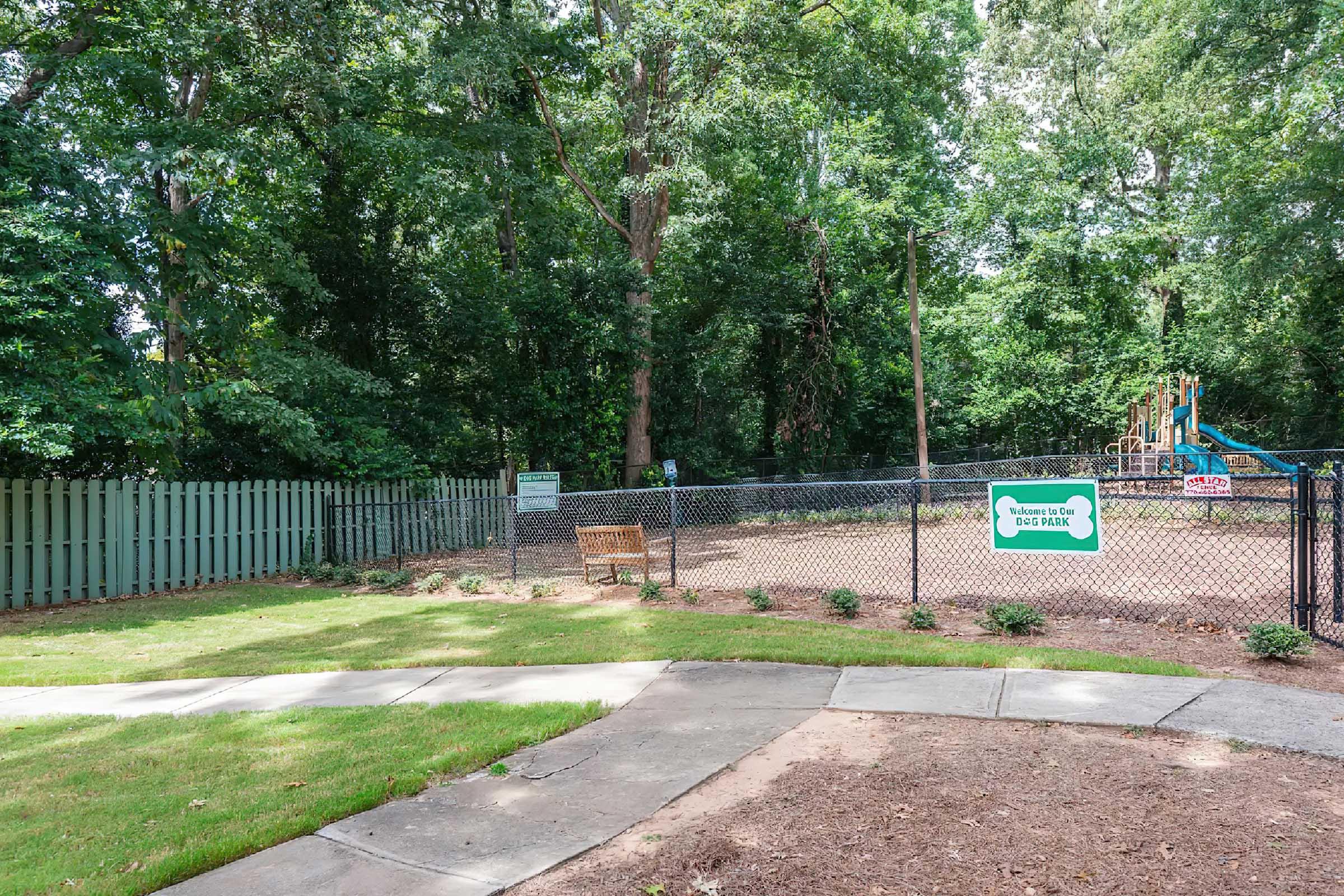 A landscaped area featuring a fenced dog park with a bench, surrounded by trees. A playground with slides is visible in the background, and signage indicates the dog park's rules. The pathway leads towards the park, and the greenery enhances the tranquil atmosphere.