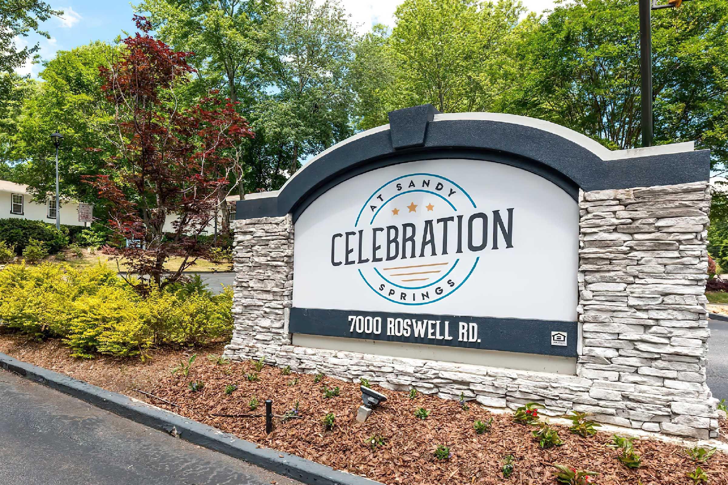 Sign for "Celebration at Sandy Springs" with the address "7000 Roswell Rd." surrounded by landscaped greenery, including small bushes and trees. The sign features a modern design with a stone base and bold lettering. Bright blue sky in the background.