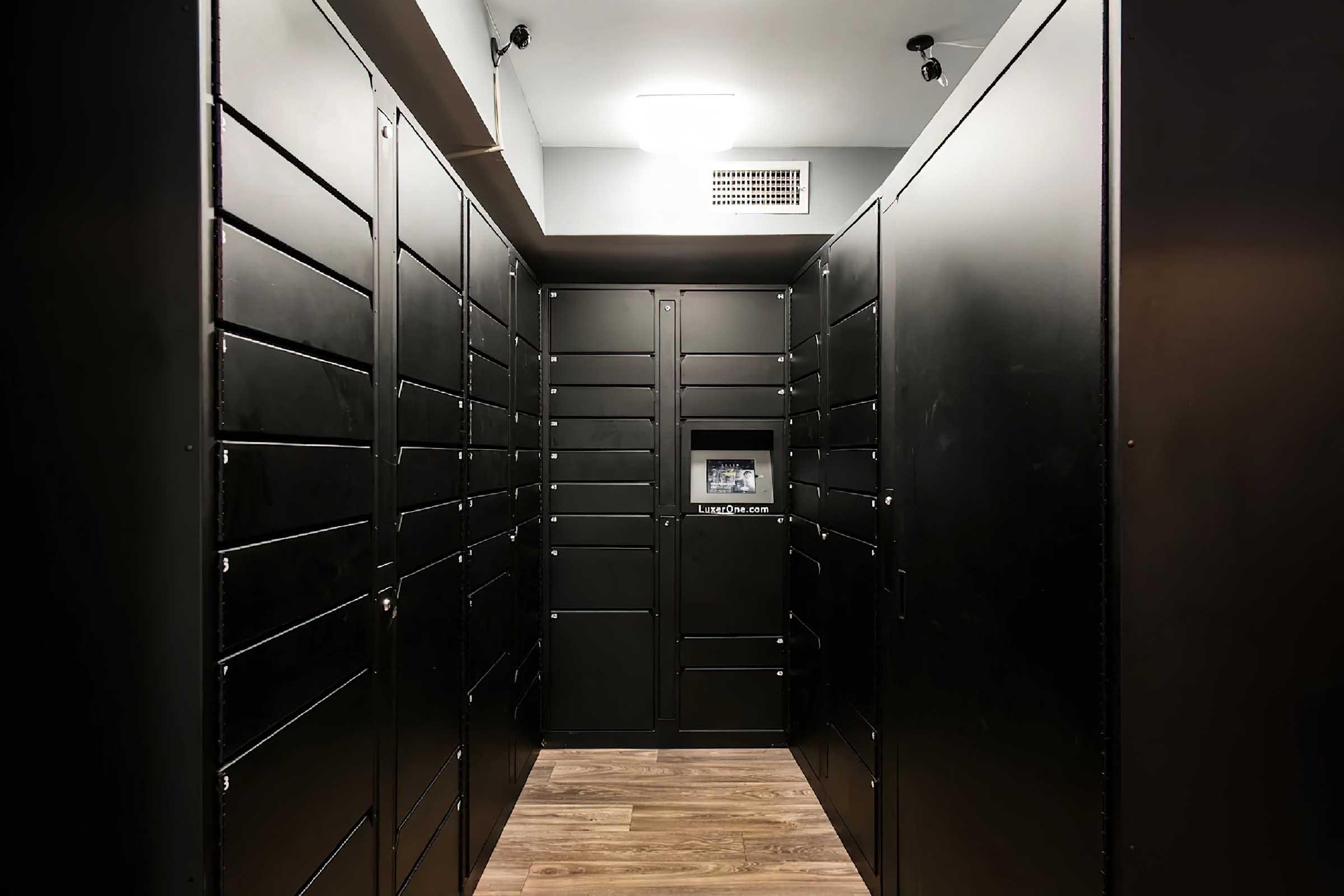 Interior of a modern package delivery room featuring black storage lockers lined on both sides with a central display unit. The floor is wooden, and the space is well-lit with overhead lighting. The atmosphere is clean and organized, designed for secure package retrieval.