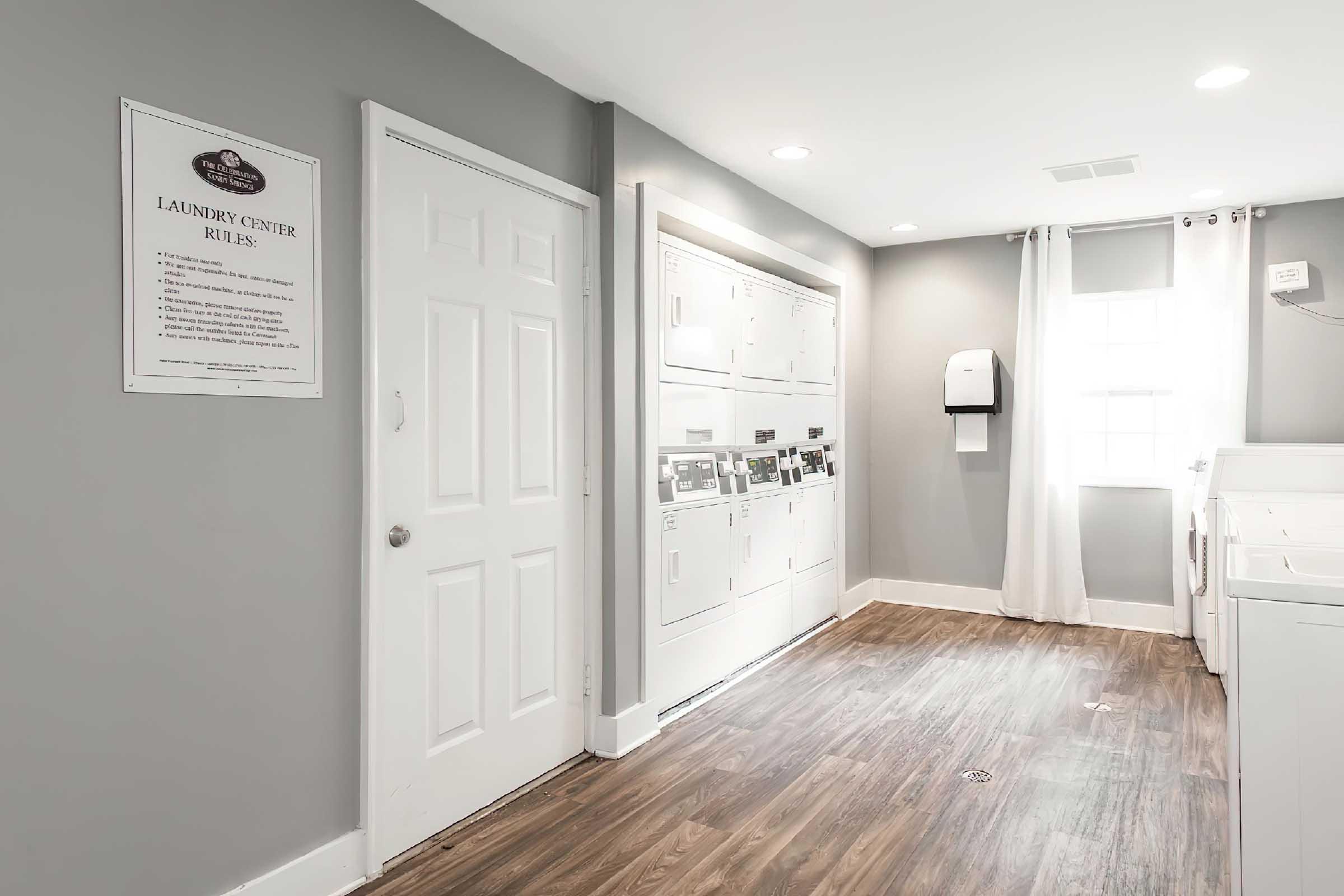 A bright and organized laundry room featuring white appliances, a wall-mounted paper towel dispenser, and a clearly visible rules sign. The space has gray walls, wood-like flooring, and a large window letting in natural light, with a door leading to another area.