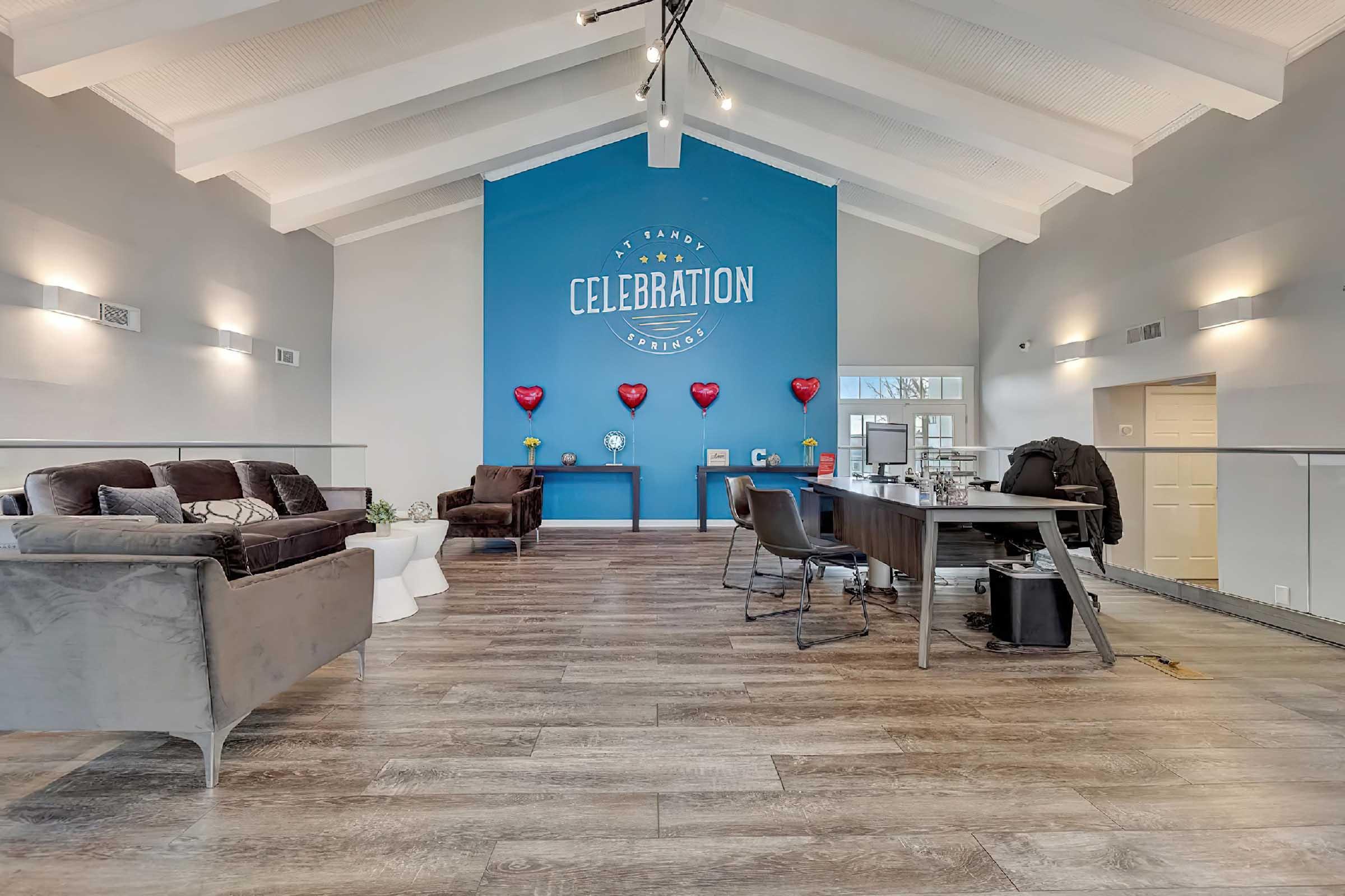 A spacious and modern interior featuring high ceilings with wooden beams. The focal point is a blue wall with the word "CELEBRATION" in white lettering. There are plush seating areas with grey sofas, a stylish desk with chairs, and decorative red heart balloons adding a festive touch to the room.