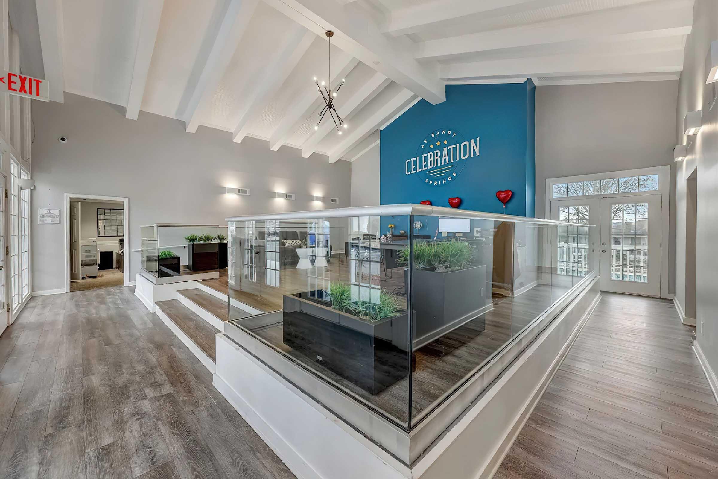 Modern interior of a lobby area featuring a bright blue accent wall with the word "CELEBRATION." The space includes glass partitions, wooden flooring, green plants in planters, and large windows allowing natural light. Stylish lighting fixtures and heart decorations add a touch of elegance.