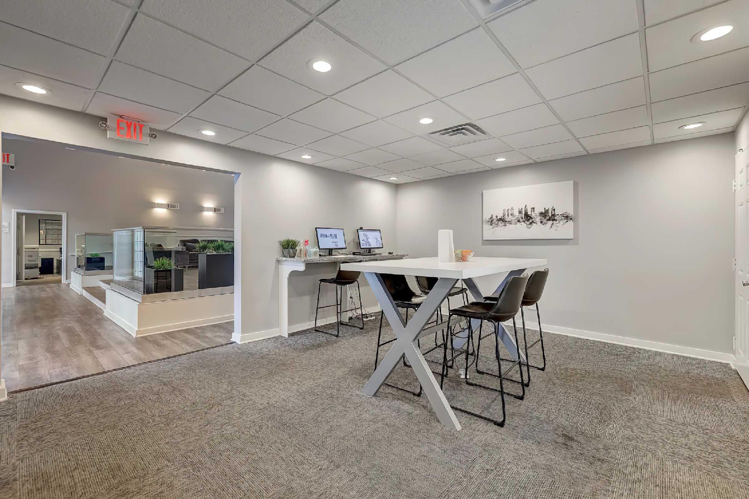 A modern office space featuring a light grey color scheme. In the foreground, there is a large white table with high-backed chairs. Two computer stations are set up against the wall with a minimalist painting above. Background includes a reception area with greenery and a stylish design. Well-lit and spacious environment.