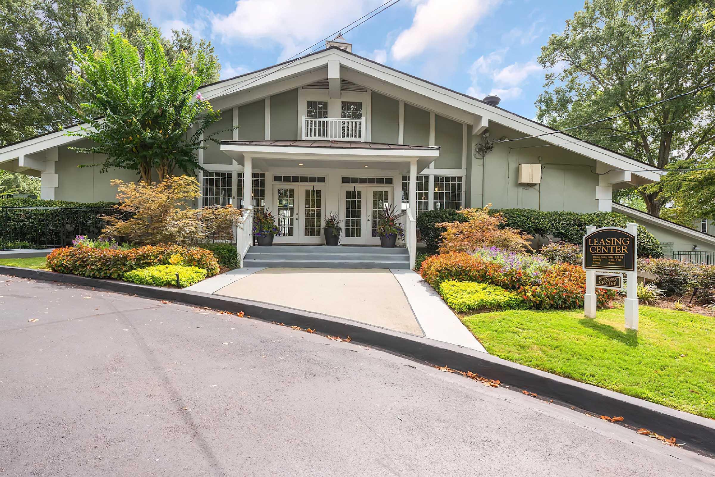 A well-maintained leasing center with a welcoming entrance, featuring a landscaped pathway leading to double doors. Surrounding the entry are colorful shrubs and trees, under a clear blue sky with fluffy clouds. A sign stating "Leasing Center" is visible near the pathway.