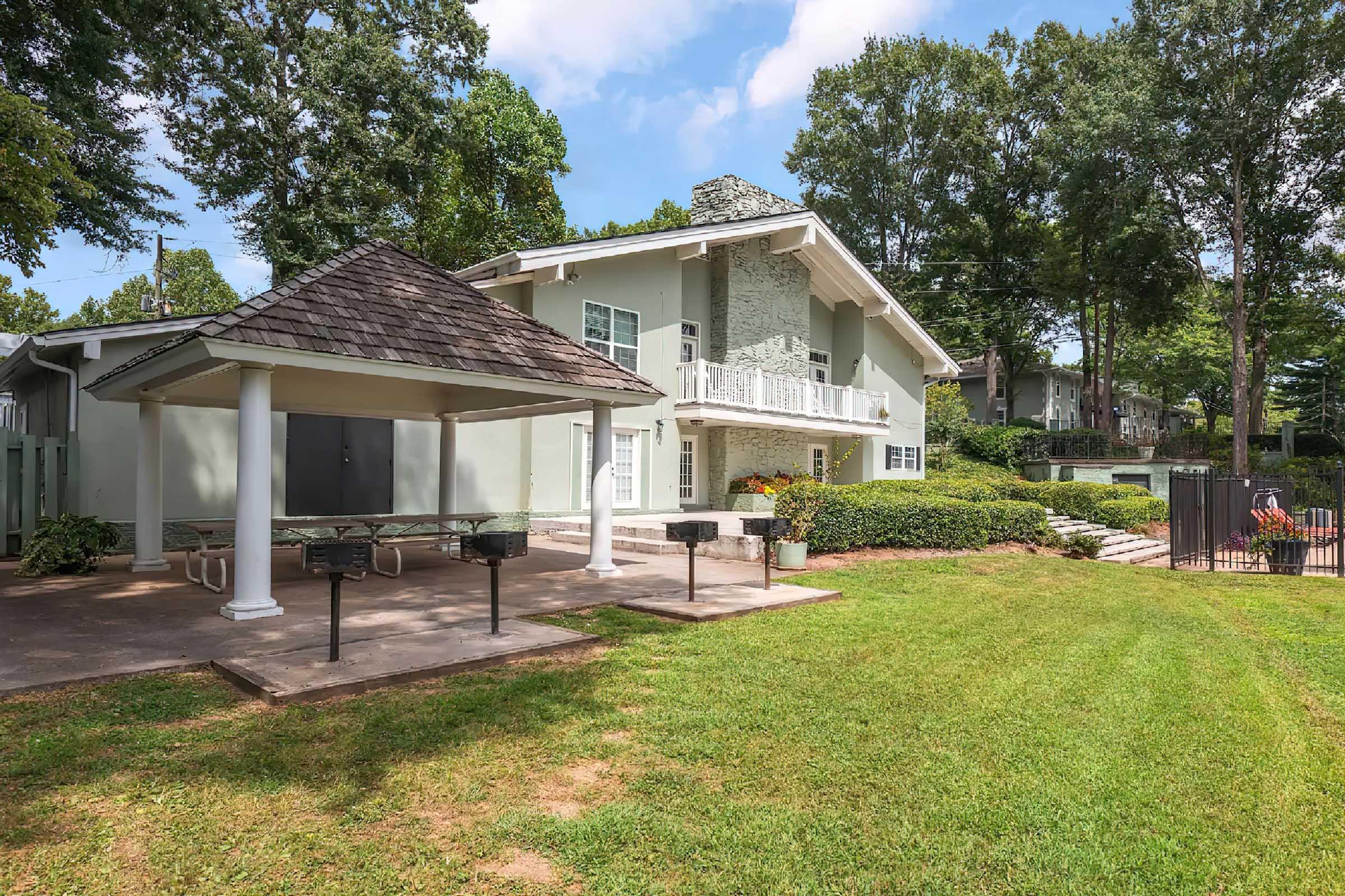 A spacious residential property featuring a two-story building with a stone chimney, surrounded by well-maintained lawns and trees. In the foreground, there is a covered picnic area with tables and a grill, and a fenced pool area is visible in the background, providing a relaxing outdoor space.