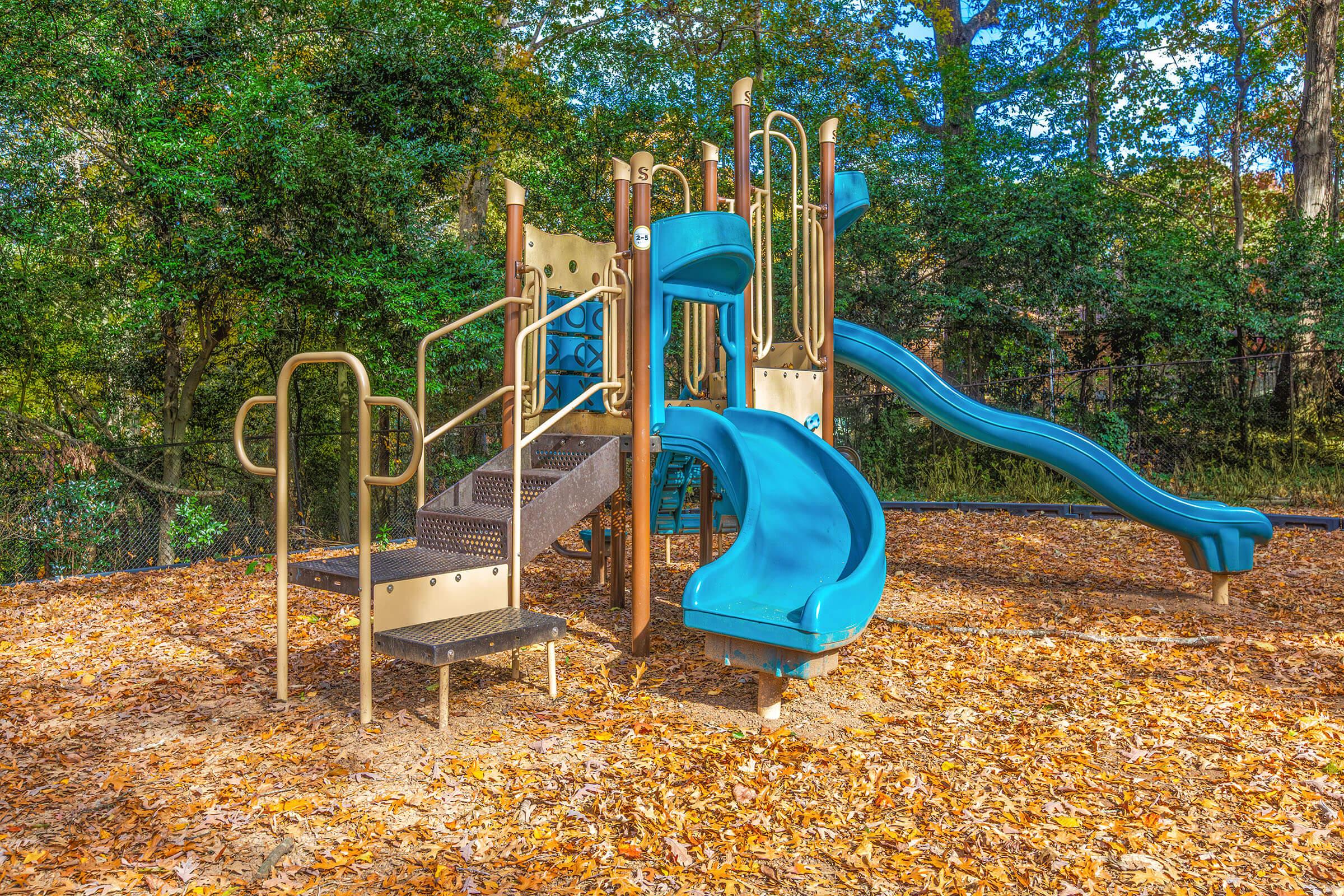 A colorful playground structure featuring multiple slides, climbing stairs, and safety rails, surrounded by a carpet of fallen leaves and bordered by green bushes and trees. The scene is bright with clear blue sky visible above.