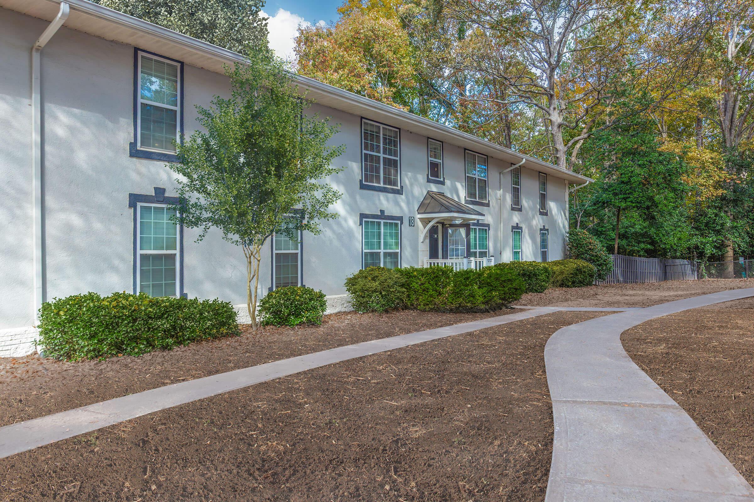 A well-maintained two-story residential building with blue shutters and a front entrance, surrounded by trimmed hedges and a freshly landscaped garden. A curved walkway leads up to the entrance, flanked by bare soil ready for planting, set against a backdrop of green trees and a clear blue sky.