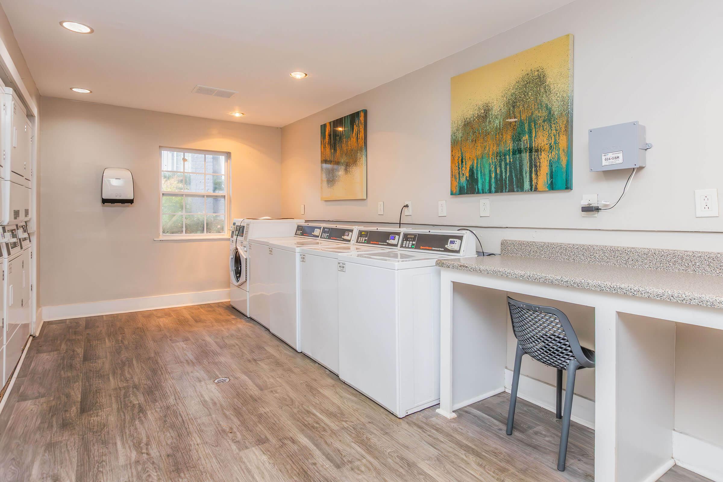 A bright and clean laundry room featuring several white washing machines and dryers, a countertop space with a chair, and colorful abstract paintings on the wall. The space is well-lit with large windows providing natural light, and the floor has a modern wood-like design.