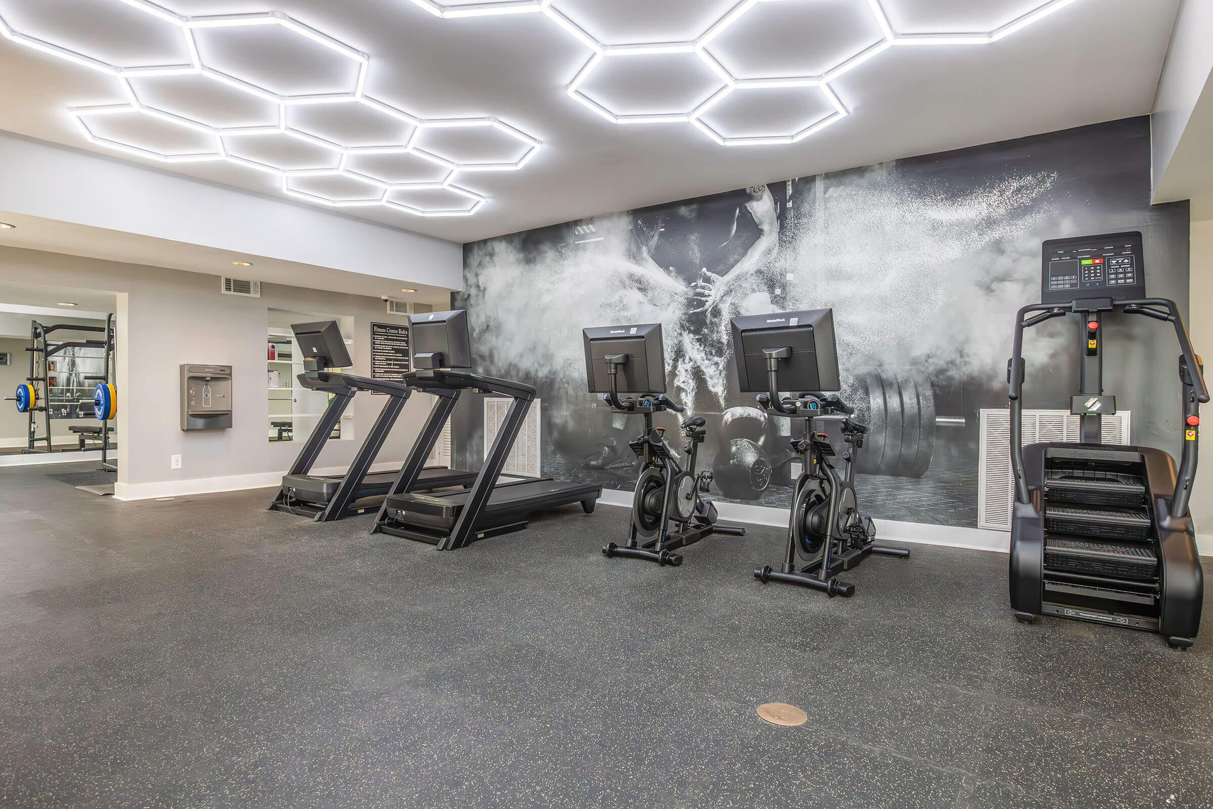 A modern gym interior featuring multiple exercise machines, including treadmills, stationary bikes, and a stair climber. The walls have a stylish design with hexagonal lighting overhead, creating a bright and inviting workout environment. Fitness equipment is neatly arranged for convenience.