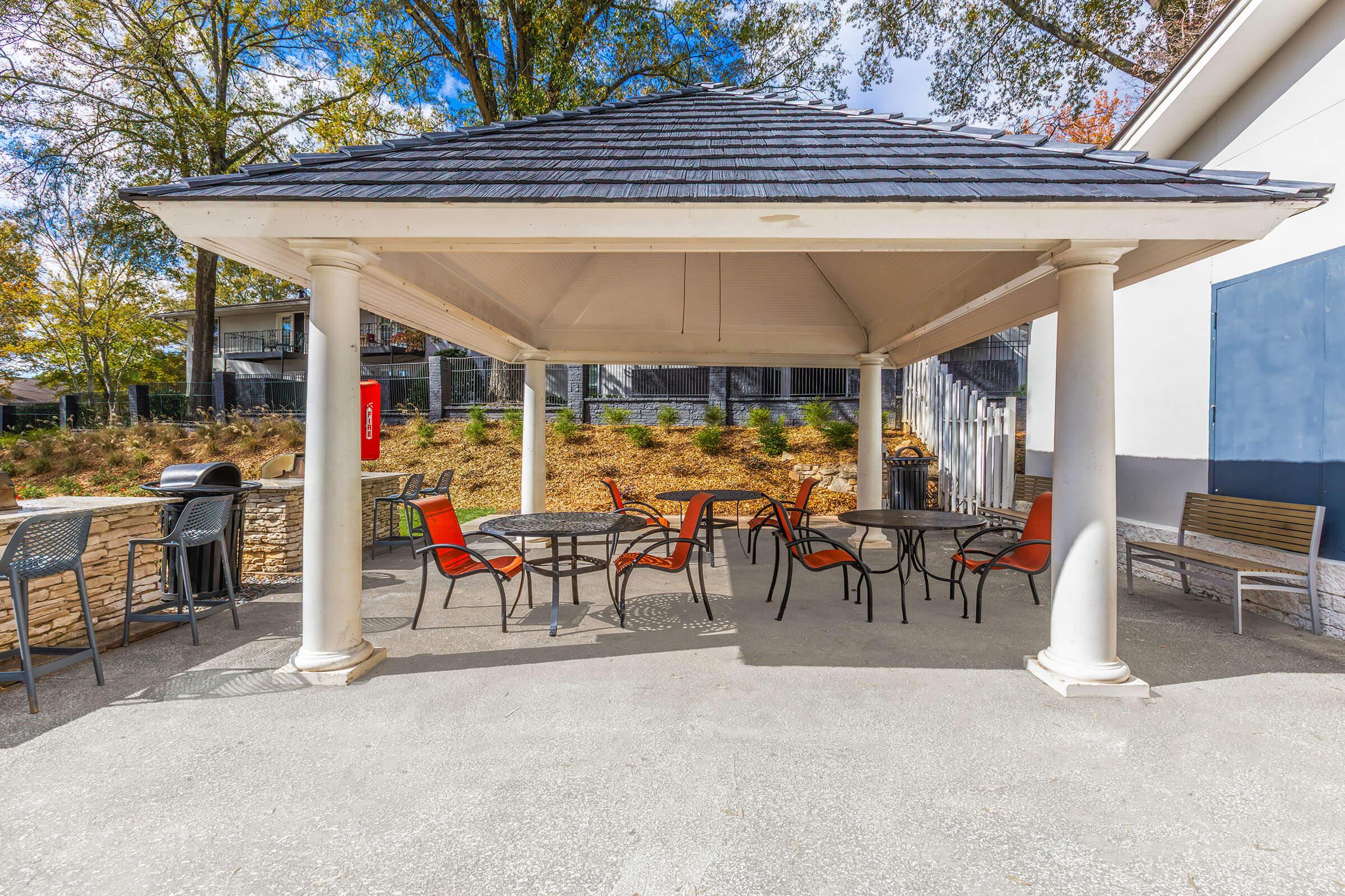 A shaded gazebo with four tables and various chairs, including orange and gray ones. The structure is surrounded by greenery and has a clean, paved floor. Visible amenities include a fire extinguisher on one column, enhancing safety in the outdoor space.