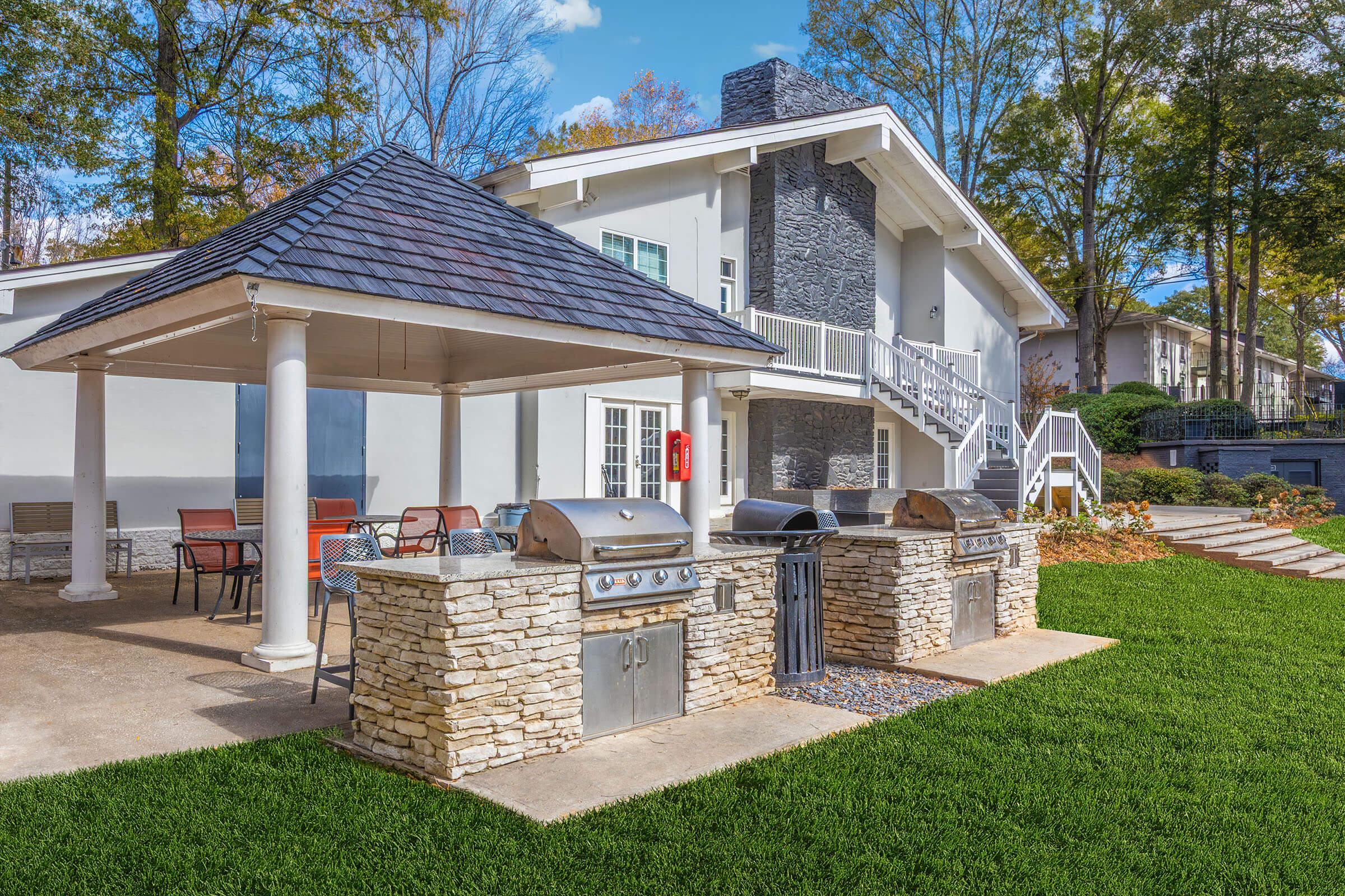 Outdoor grilling area featuring two stone BBQ grills under a gazebo, surrounded by patio seating. The background showcases a modern house with a gray stone wall, lush green lawn, and trees. Clear blue skies with a few clouds complete the inviting atmosphere.