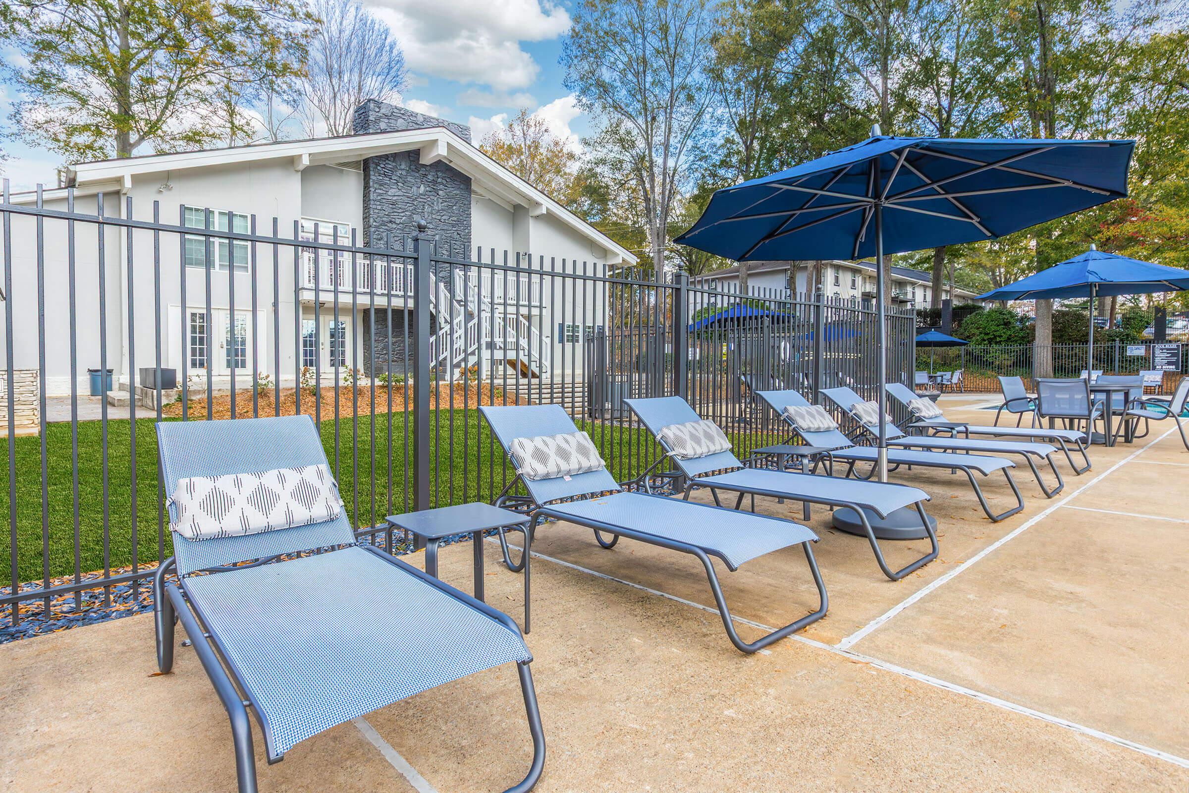 Lounge chairs arranged by a swimming pool area, with blue umbrellas providing shade. A grassy lawn and a two-story building are visible in the background under a partly cloudy sky. The setting appears clean and inviting, ideal for relaxation.