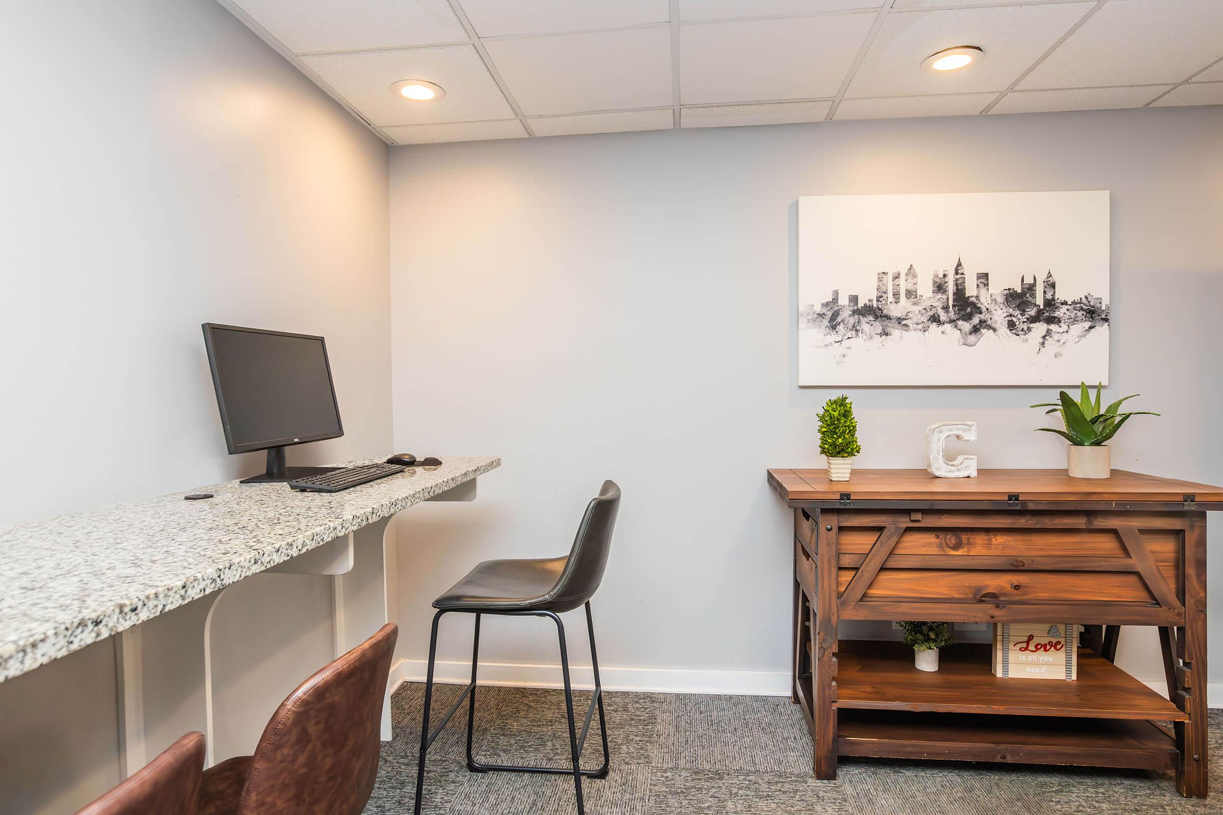 A small office space featuring a granite countertop desk with a computer monitor, a black chair, and a wooden table. The wall is decorated with a framed cityscape artwork, and decorative plants are placed on the table. The room has neutral-colored walls and overhead lighting.