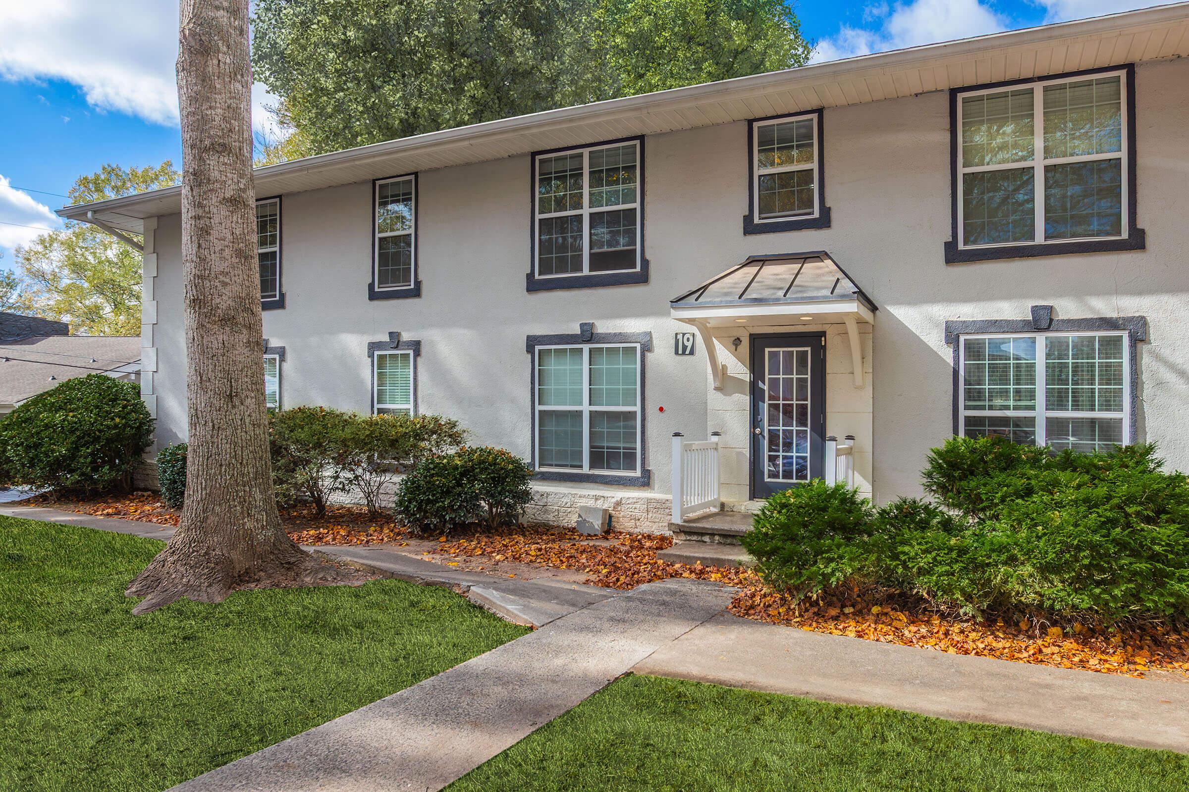 A two-story residential building featuring a front porch with a steps entry, surrounded by lush green landscaping and autumn leaves scattered on the pathway. The facade has multiple windows framed in black, and the building is situated in a serene environment with trees nearby.