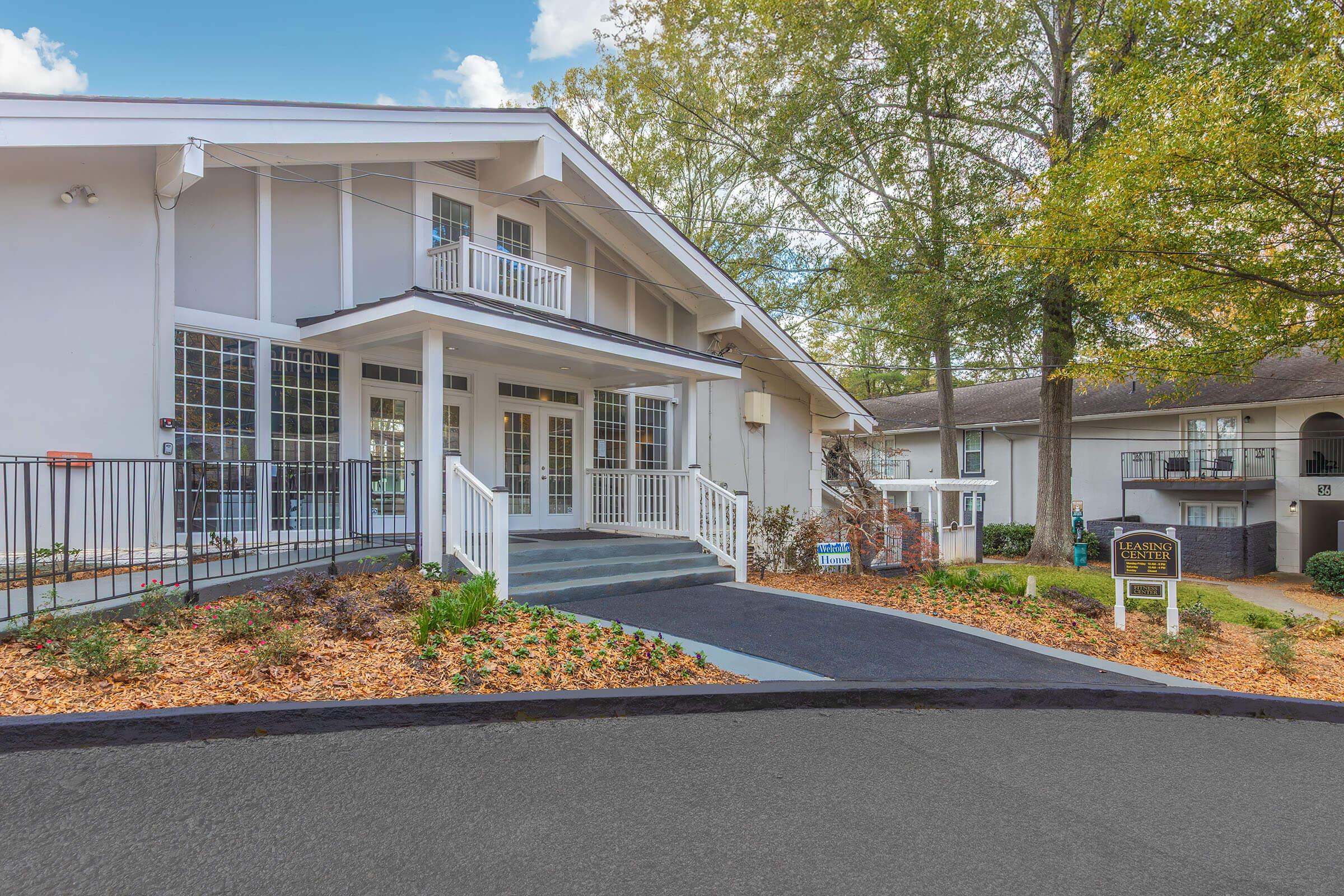 A modern building entrance with a welcoming porch, surrounded by landscaped flower beds and small trees. The facade features large windows and a sign indicating the location. Six steps lead up to the main door, nestled between a parking area and additional structures in the background.