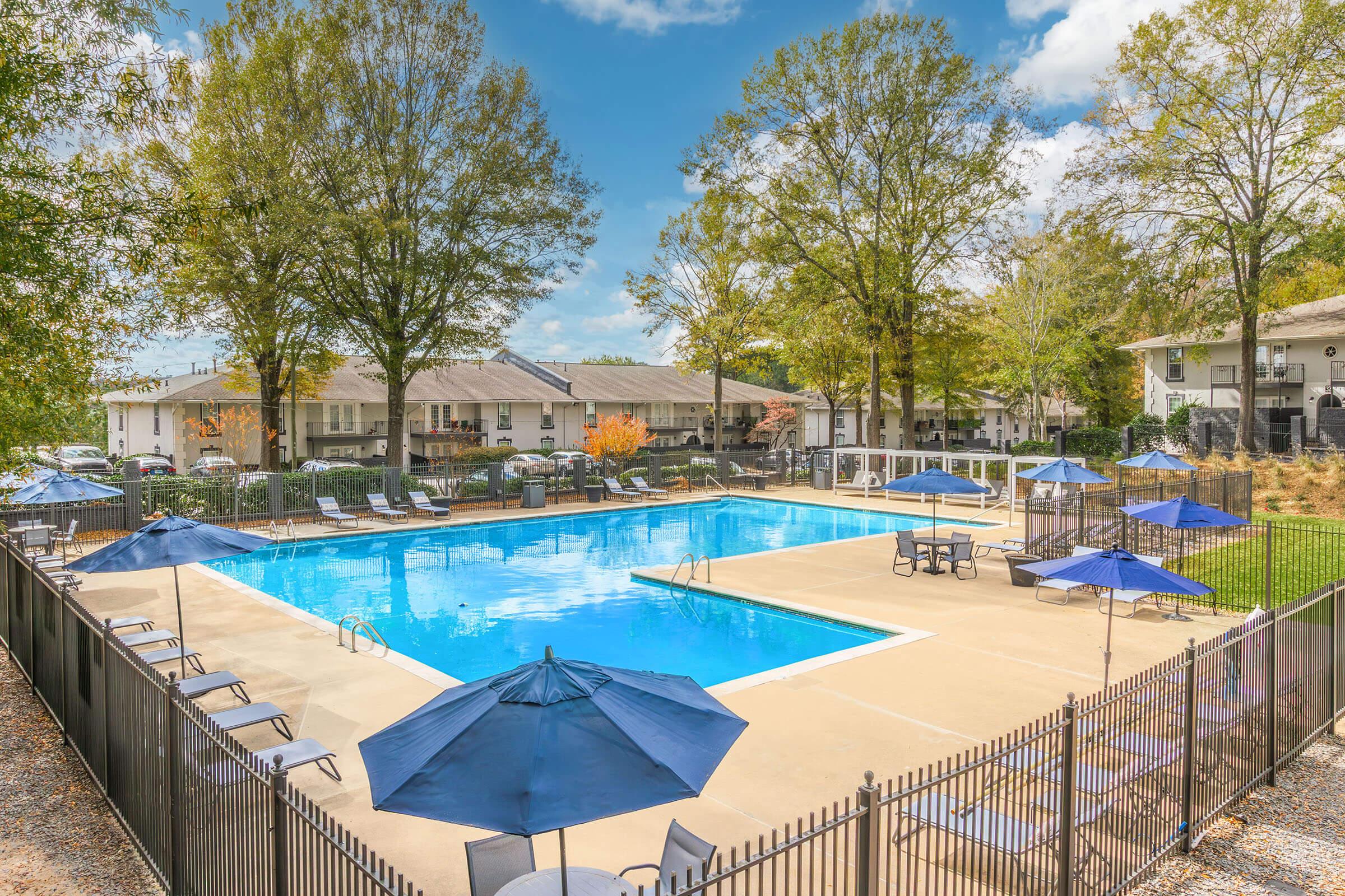 A spacious outdoor swimming pool surrounded by lounge chairs and umbrellas. The pool area is bordered by a black fence and features trees in the background, along with several apartment buildings. Bright blue skies and fluffy clouds enhance the inviting atmosphere of the scene.