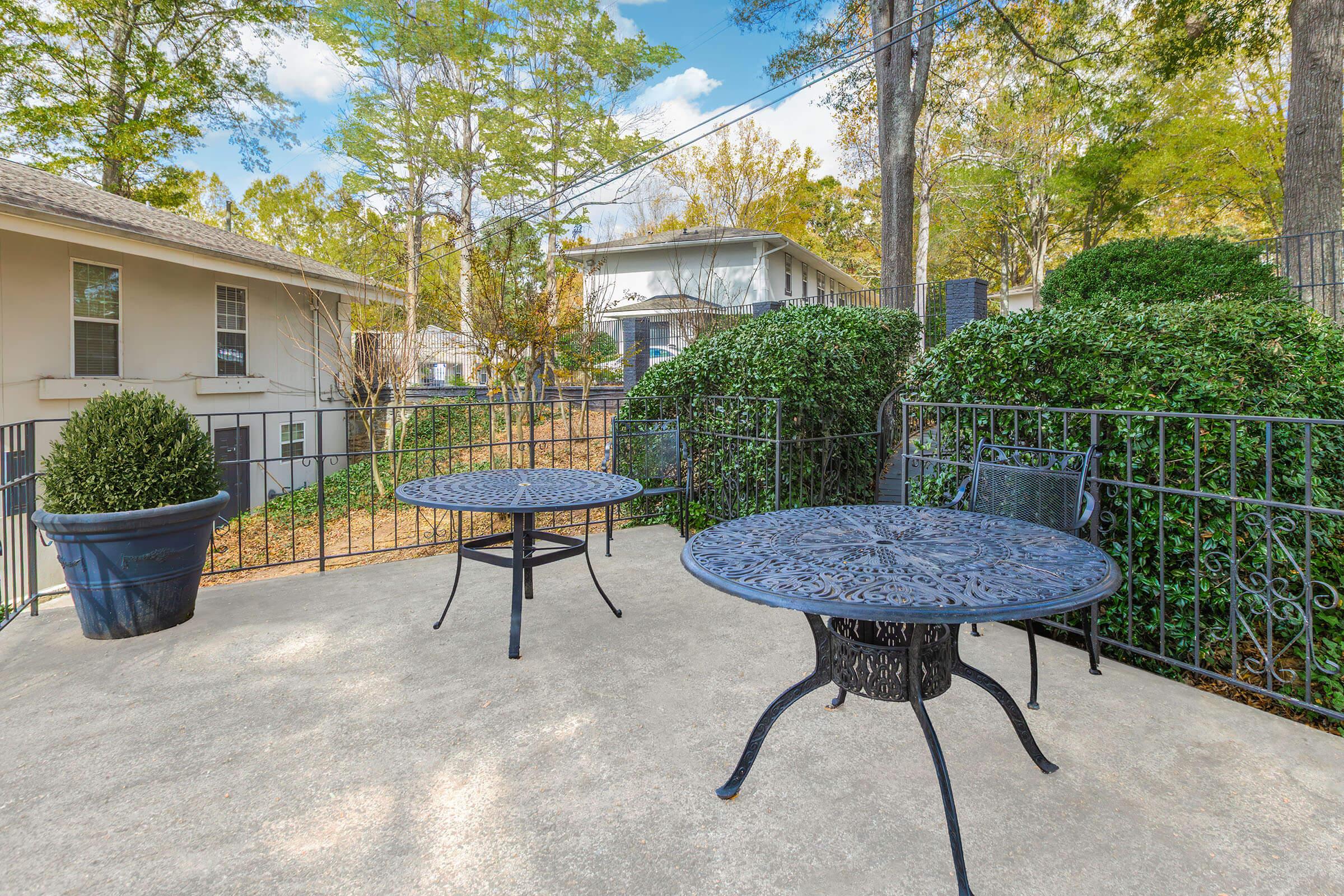 Two wrought iron tables with chairs on a concrete patio, surrounded by neatly trimmed hedges and greenery. In the background, a glimpse of two buildings and trees under a clear blue sky. The peaceful outdoor space is suitable for relaxation or dining.