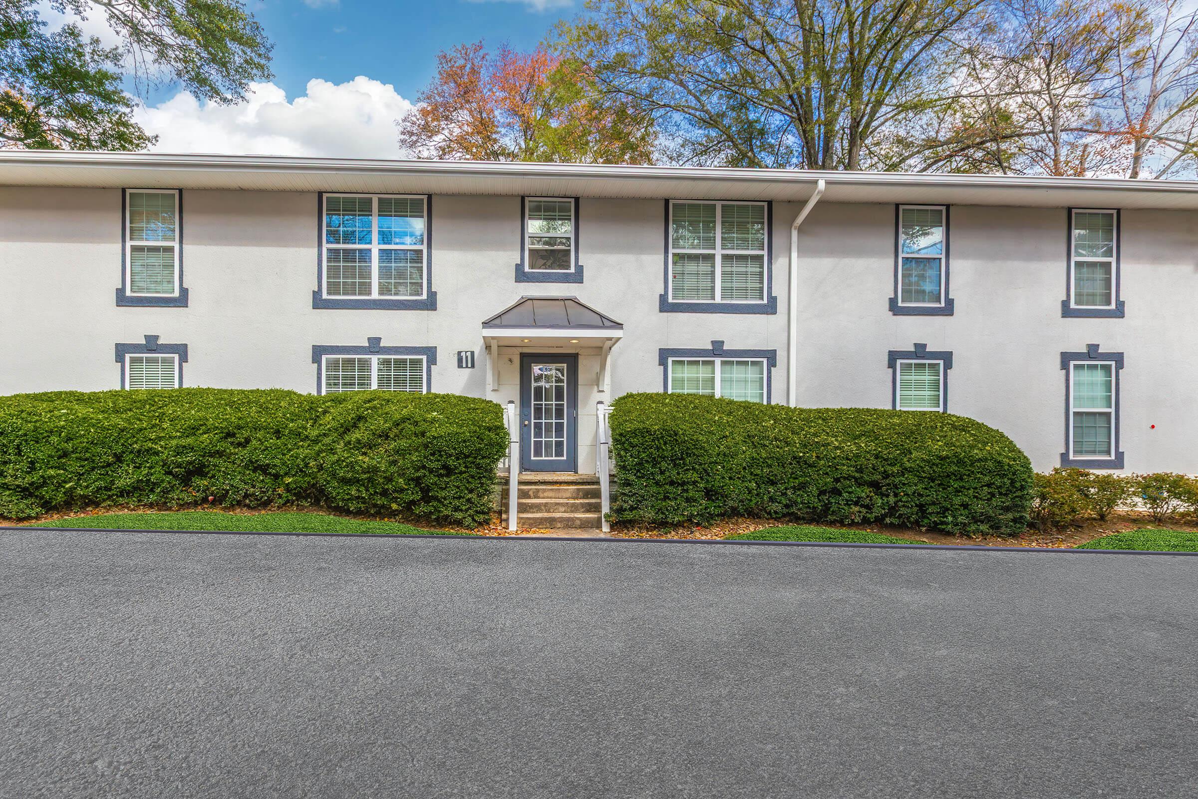 A two-story residential building with white exterior walls and dark trim. The front features several large windows, a double-door entrance, and well-manicured hedges. The driveway is paved, leading up to the entrance, with lush greenery surrounding the property under a clear blue sky.