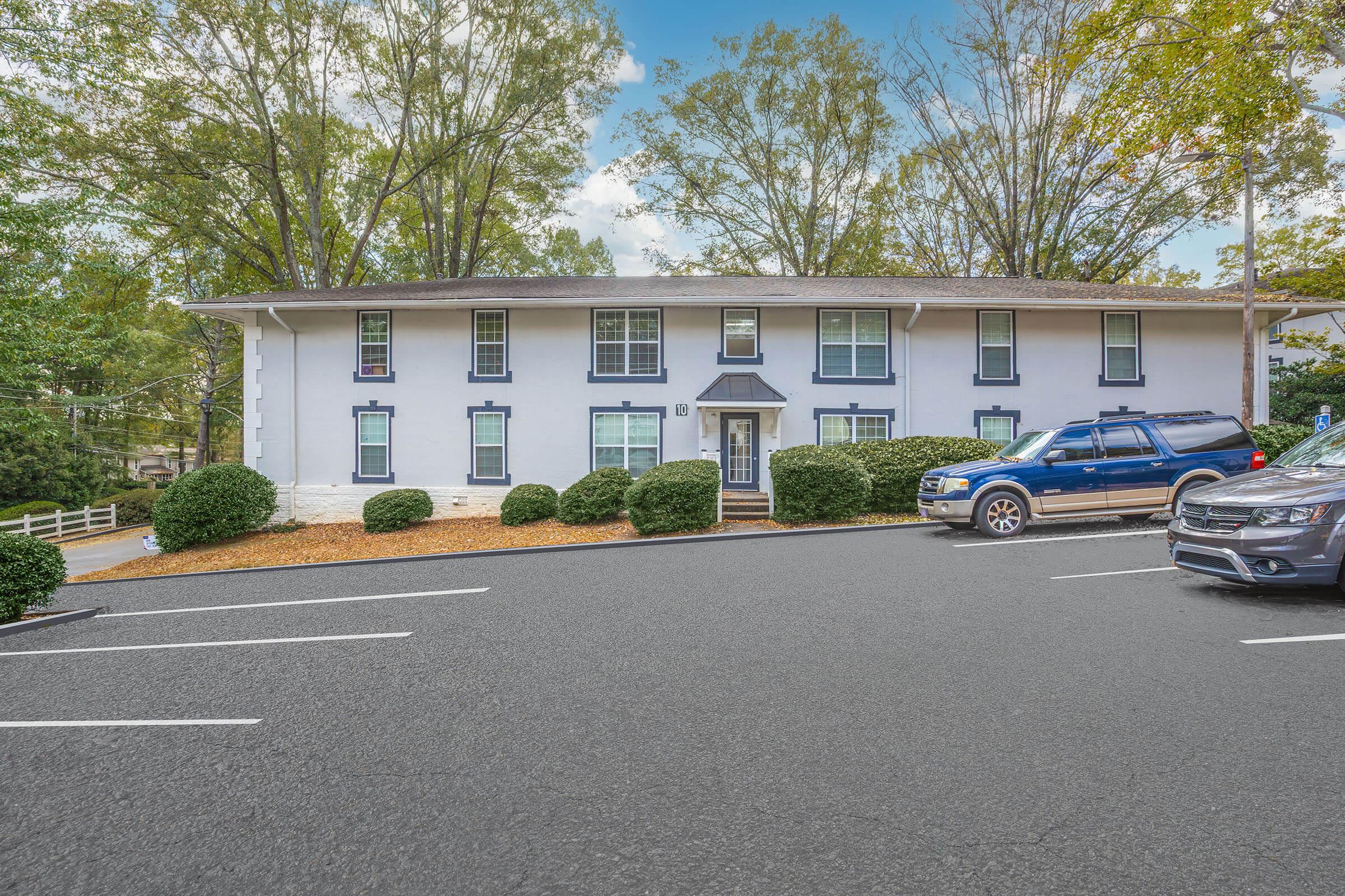 A two-story white apartment building with green shutters and a manicured front lawn. The entrance is framed by shrubs, and there are several parked cars in the foreground on a paved lot. Trees provide shade, and the sky is partly cloudy.