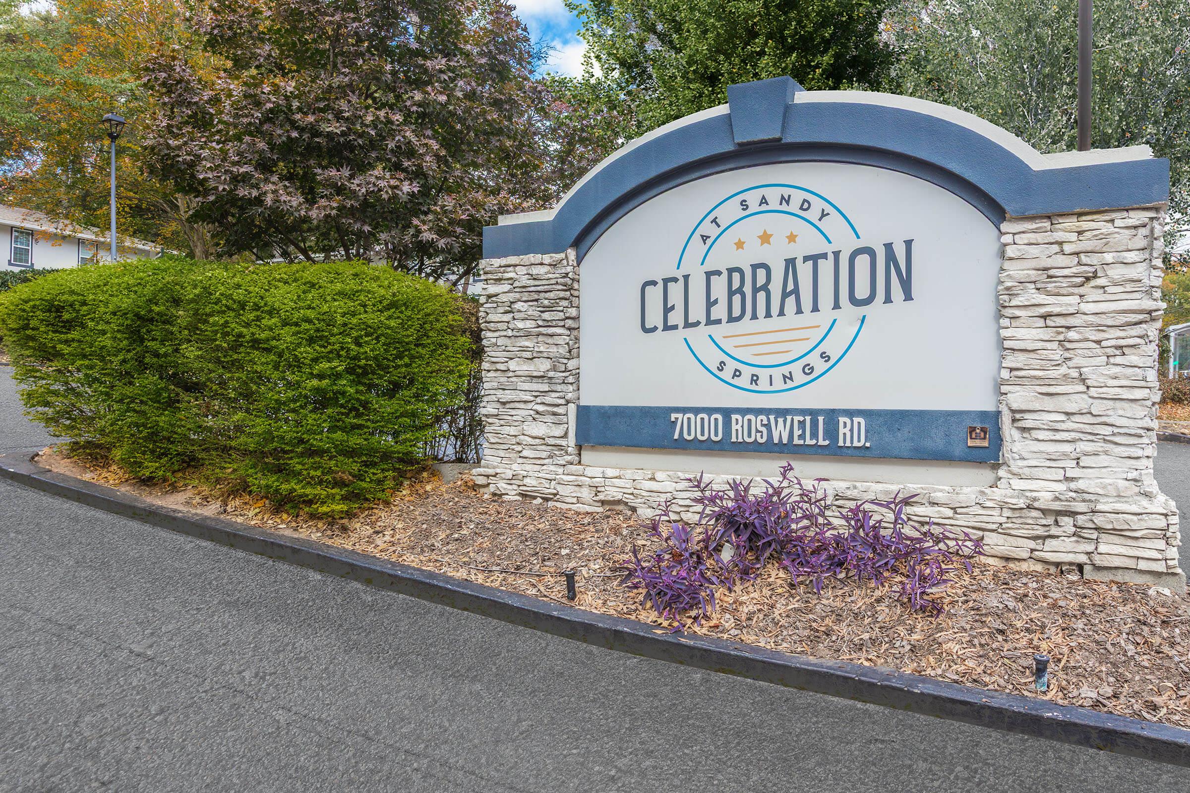 Sign for Celebration at Sandy Springs, featuring the name "Celebration" prominently in blue letters. The sign includes a stone surround and is located at the address 7000 Roswell Rd. Surrounding the sign are neatly trimmed green bushes and decorative landscaping.