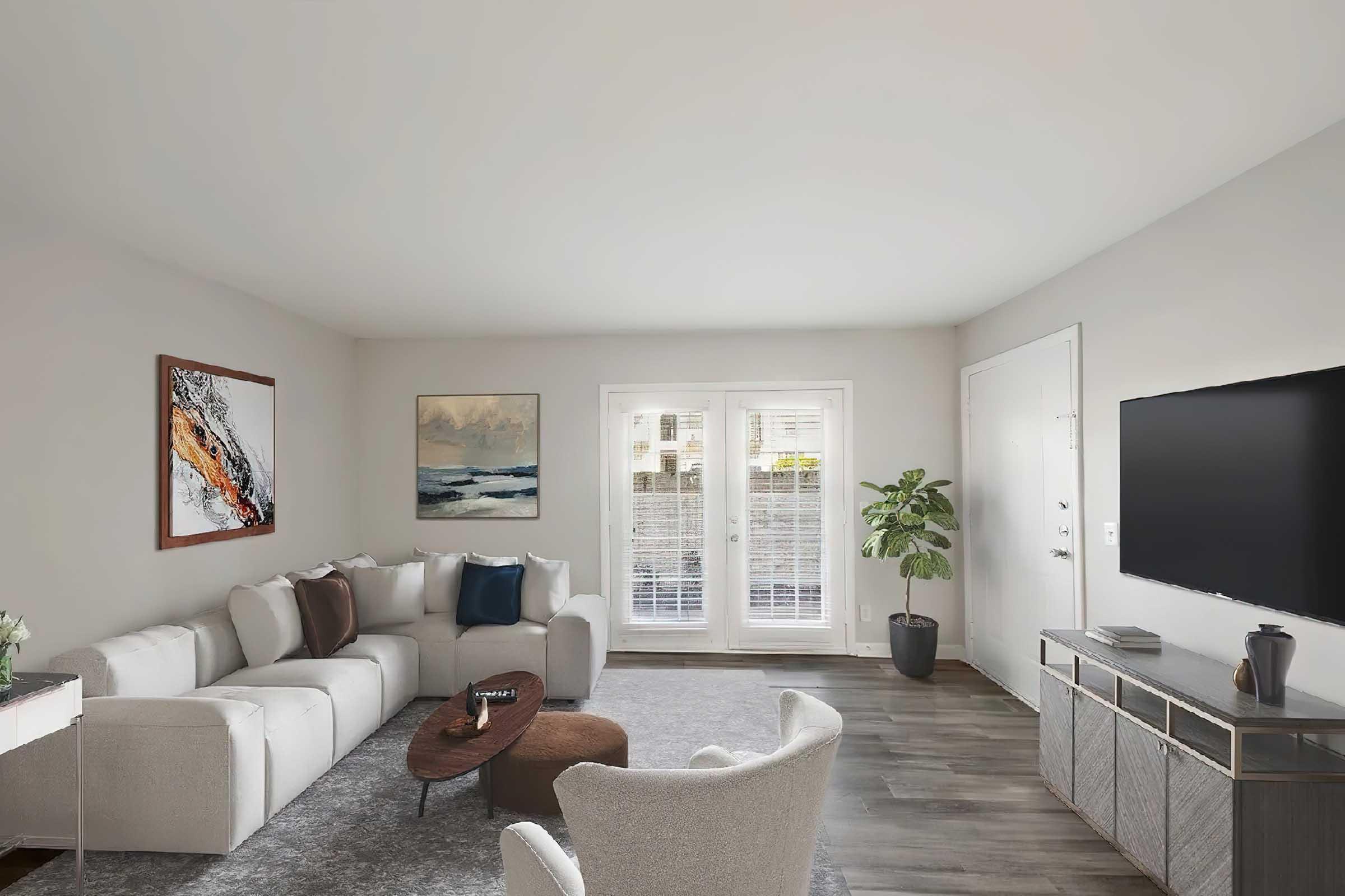 A modern living room featuring a plush white sectional sofa, a round coffee table, and a cozy accent chair. Walls are painted in neutral tones, adorned with abstract artwork. Large sliding glass doors provide natural light, leading to an outdoor space. A flat-screen TV is mounted on a stylish media console.