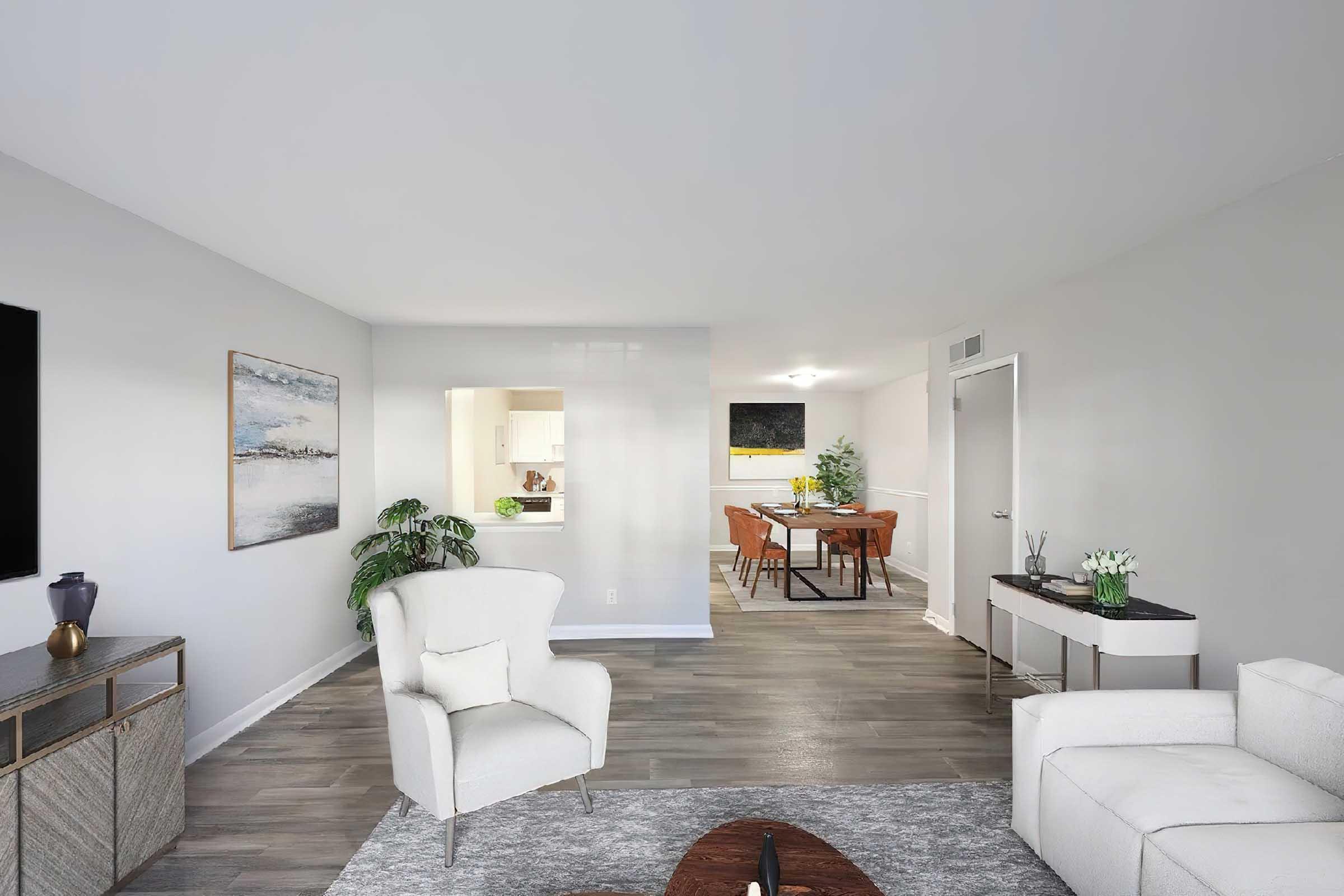 A modern living room featuring a light-colored armchair and couch, a decorative coffee table, and a textured area rug. A television is mounted on the wall, and a small potted plant adds greenery. In the background, a dining area with wooden chairs and a table is visible, along with an open kitchen space.