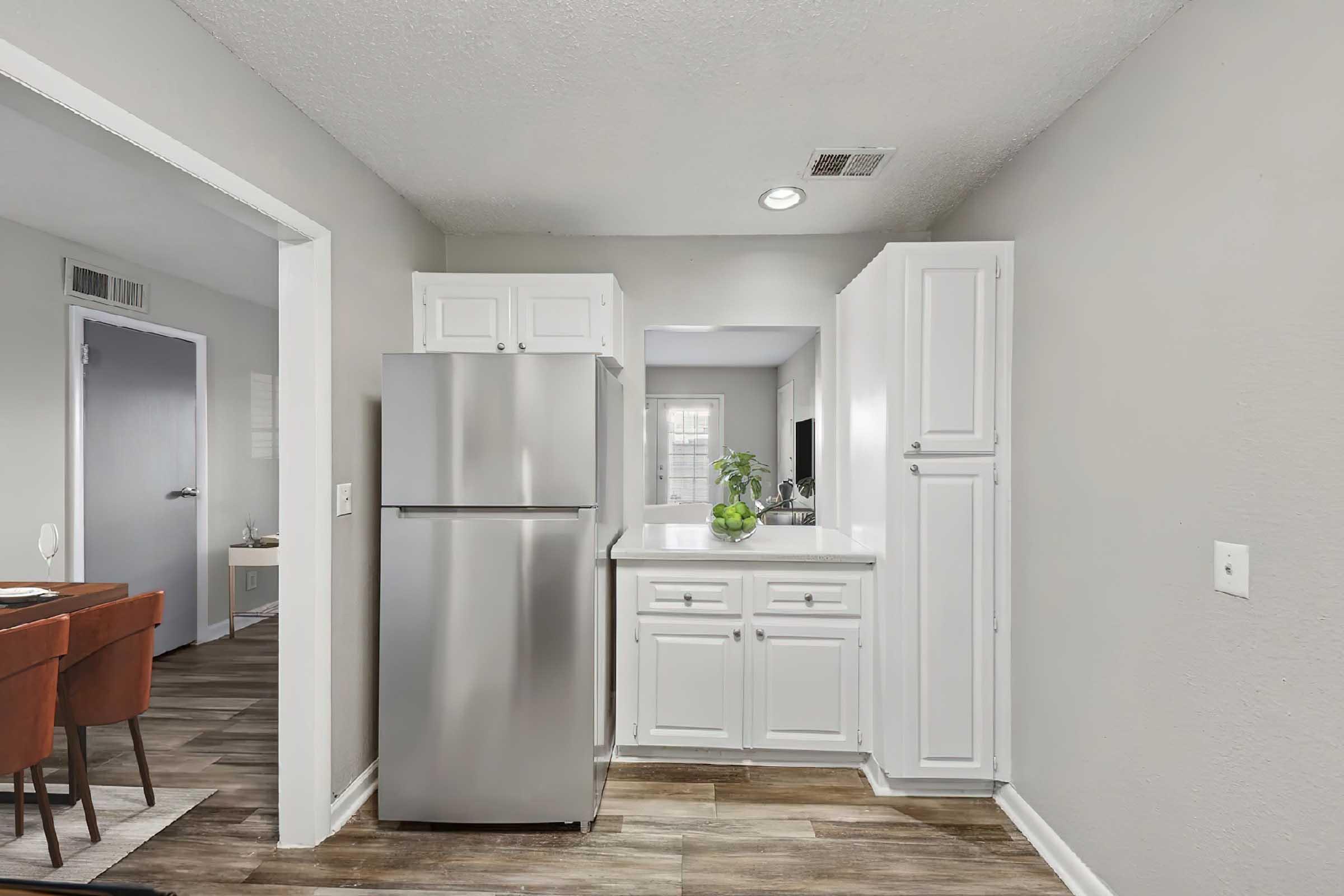 A modern kitchen featuring stainless steel appliances, including a refrigerator, with white cabinetry and a countertop. The room has a neutral color scheme and hardwood-like flooring, leading to a dining area visible through an open doorway. A small plant adds a touch of greenery to the space.