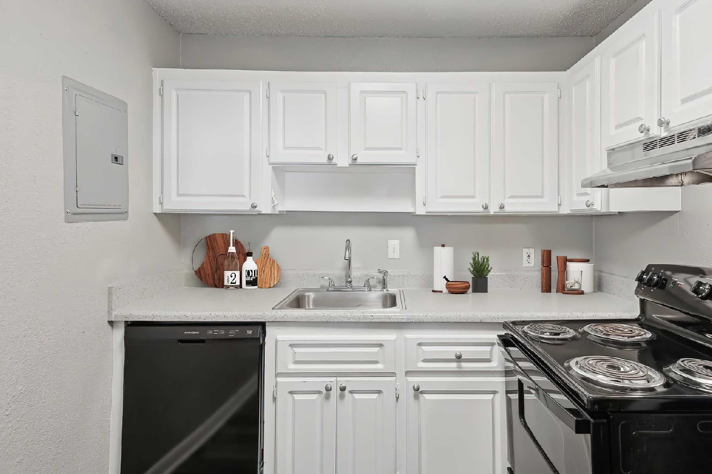 A modern kitchen featuring white cabinetry, a stainless steel sink, and a black stove. The countertop is light-colored with a wooden cutting board and decorative elements like a plant and kitchen essentials. A dark dishwasher is also visible. The overall design is clean and functional.
