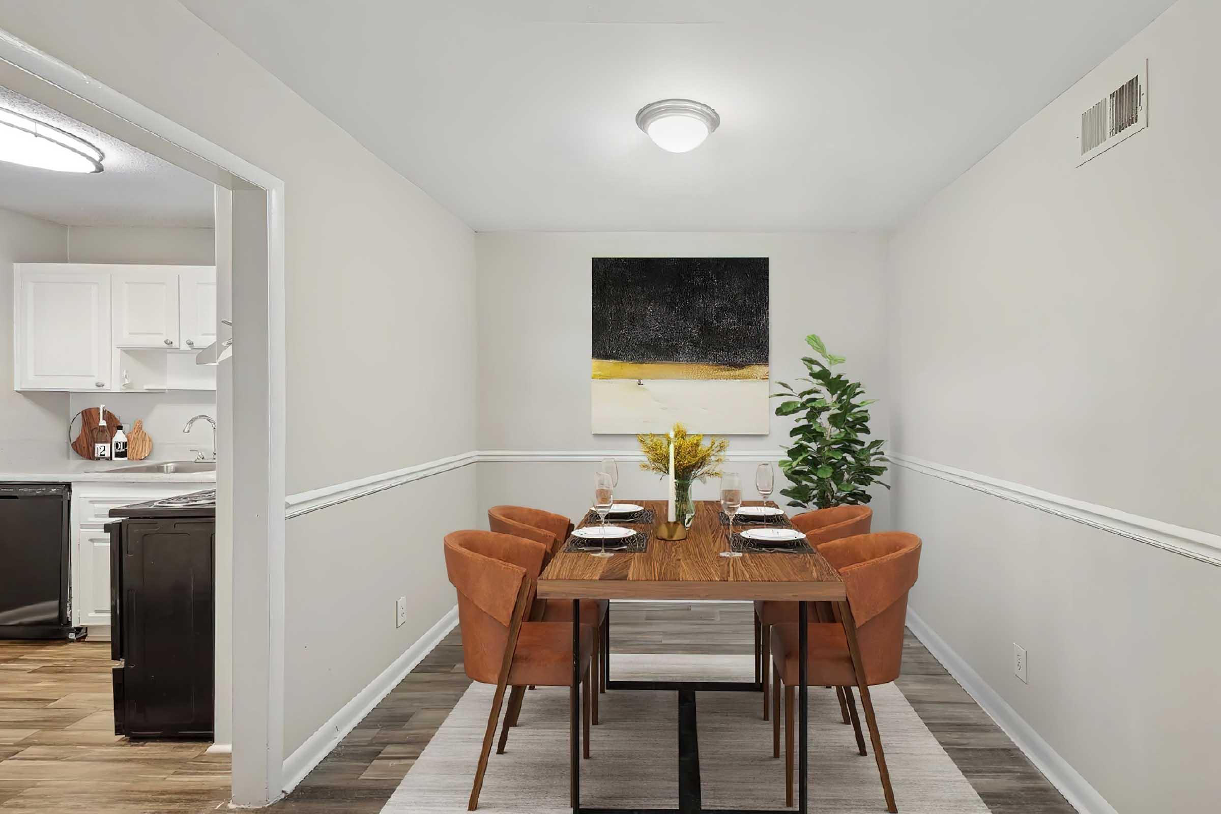 A modern dining area featuring a wooden table set with four orange chairs. A simple floral centerpiece sits on the table. The walls are painted light gray, contrasting with a large abstract painting above the table. A plant is placed in the corner, and a doorway leads into a kitchen area visible in the background.