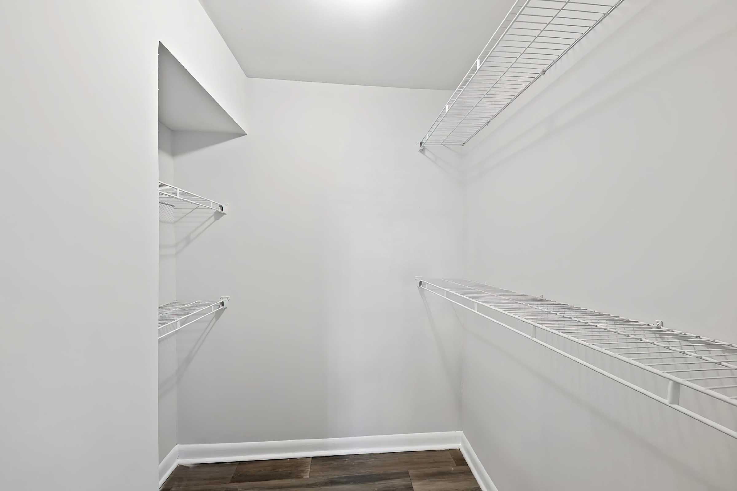 Empty closet with white wire shelving on two walls. The walls are painted a light color, and the floor is a dark wood-like surface. A small nook is visible at the back, creating an open and spacious feel.
